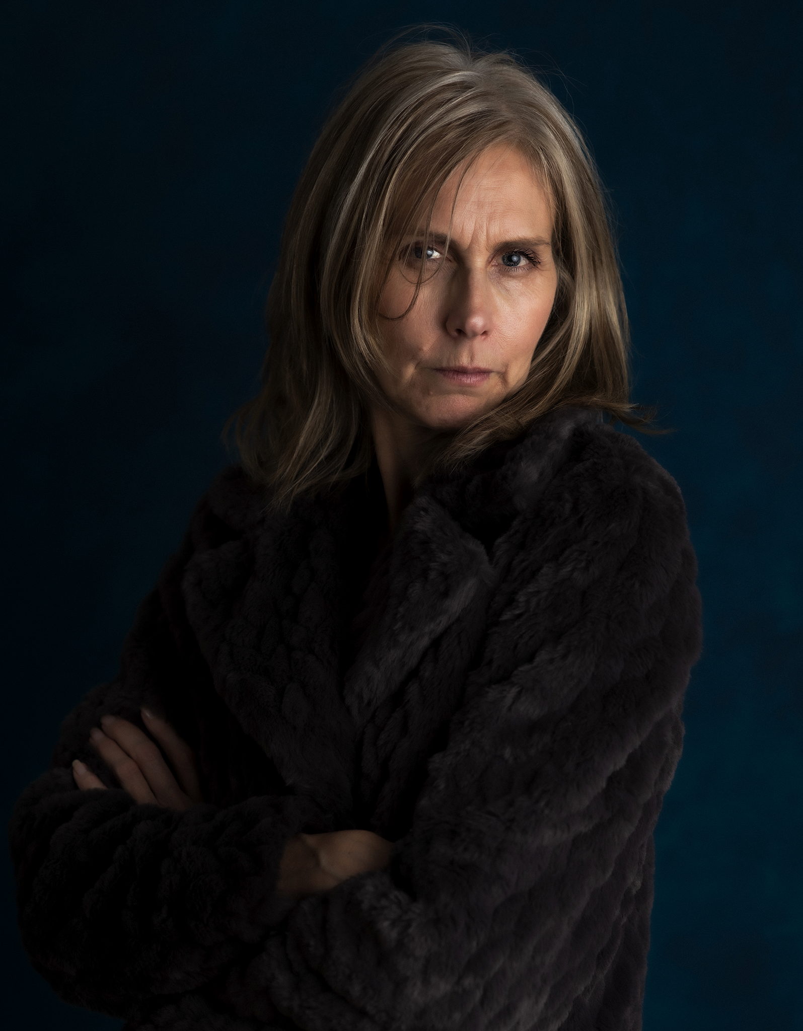 A middle-aged woman with shoulder-length blonde hair, wearing a black furry coat, looking directly at the camera with a serious expression, against a dark blue background.