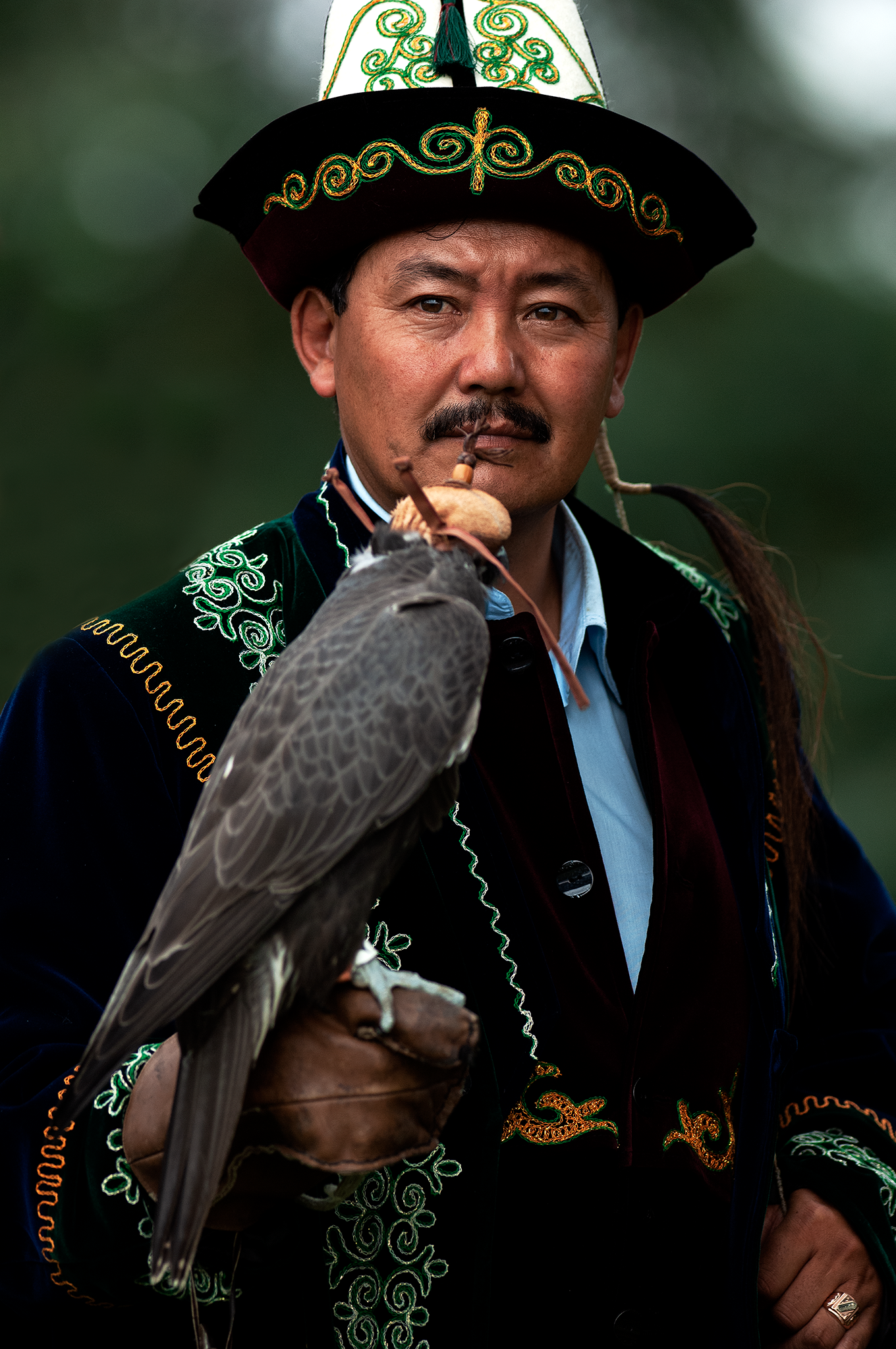 Man dressed in traditional embroidered clothing and hat, holding a falcon with a leather glove, outdoors with blurred green background.