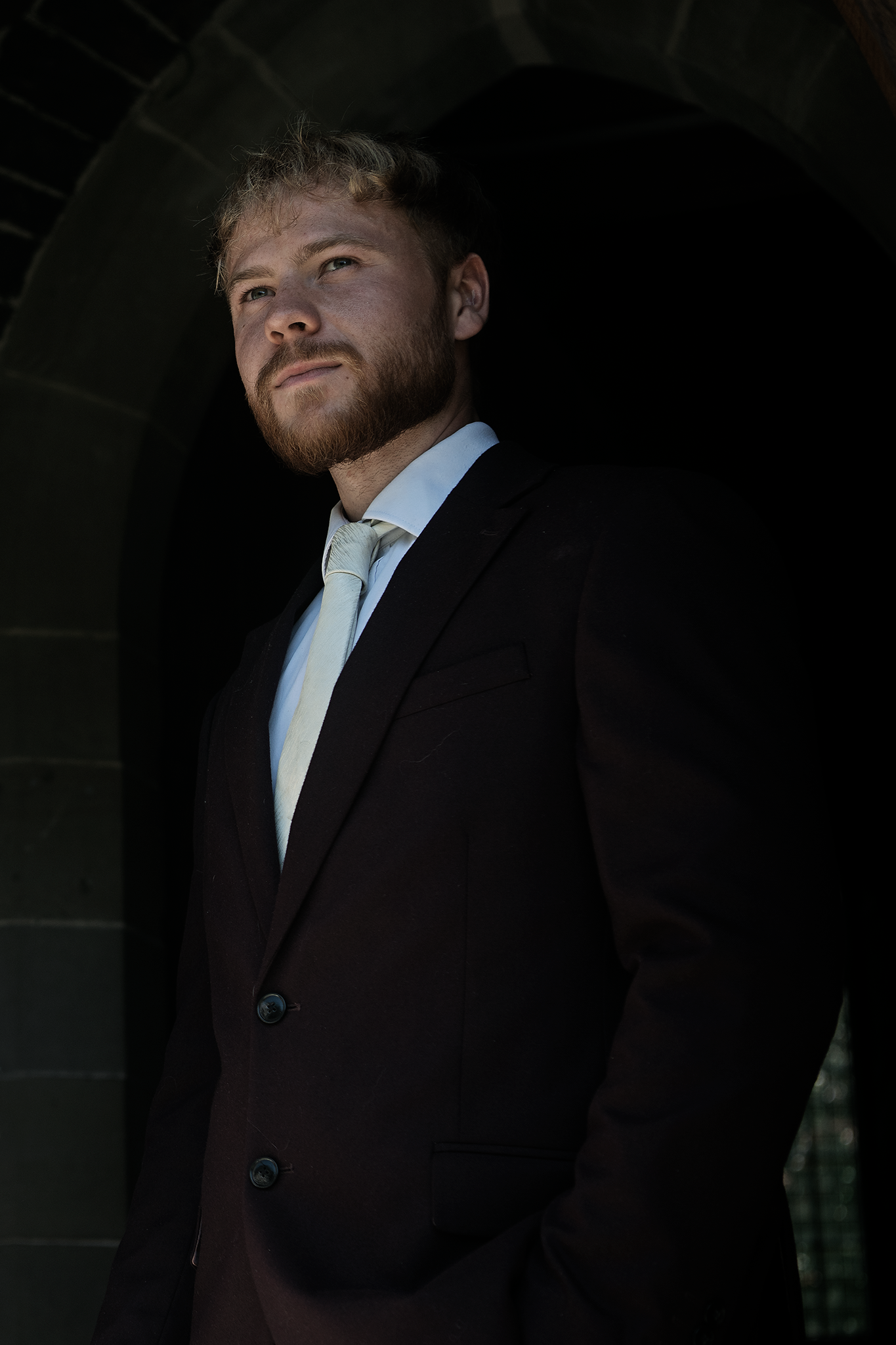 Young man with a beard wearing a black suit and a light-colored tie, standing under a dark archway.