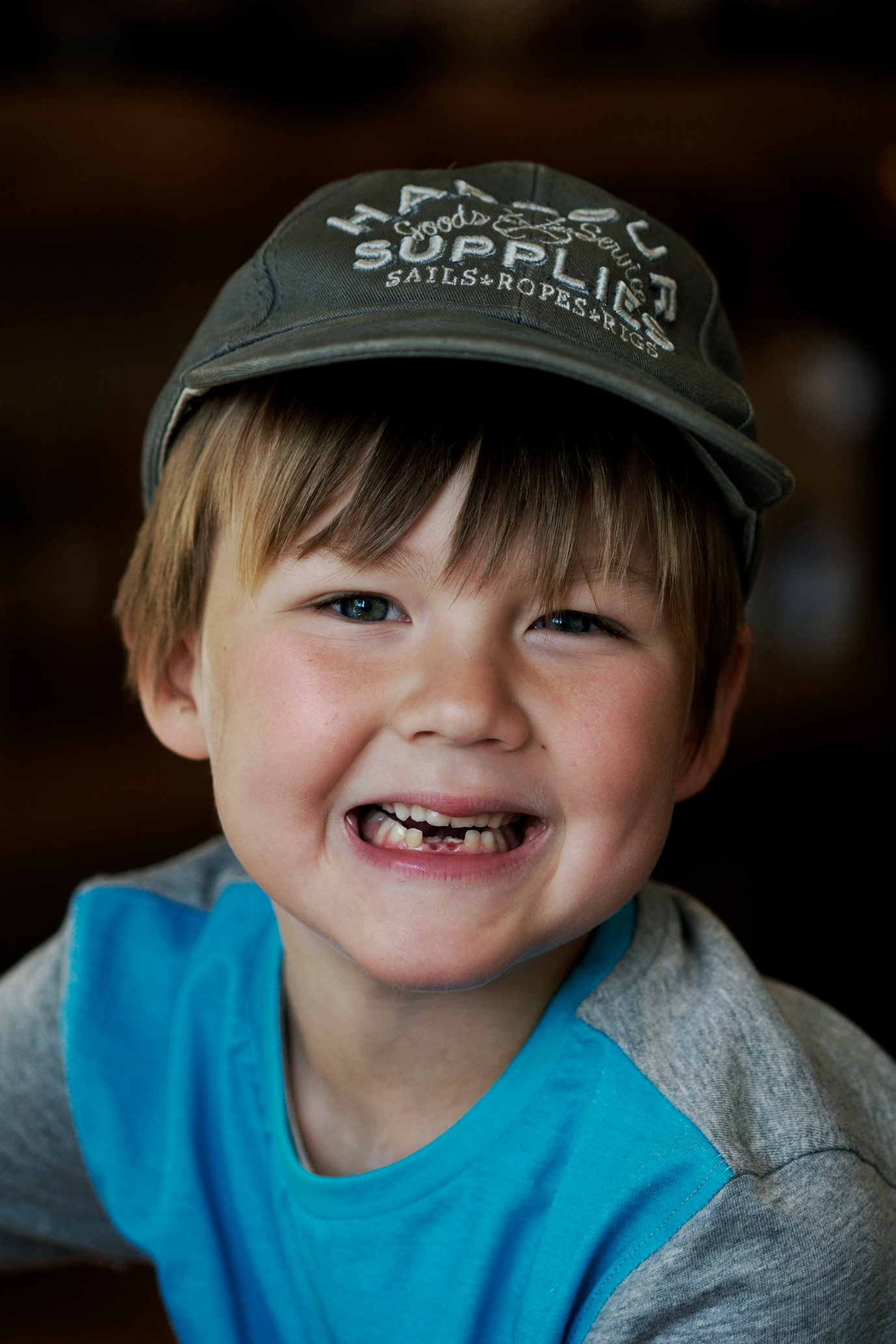 A young boy with blue eyes and a missing tooth, smiling, wearing a gray cap with white embroidered text and a blue and gray shirt.