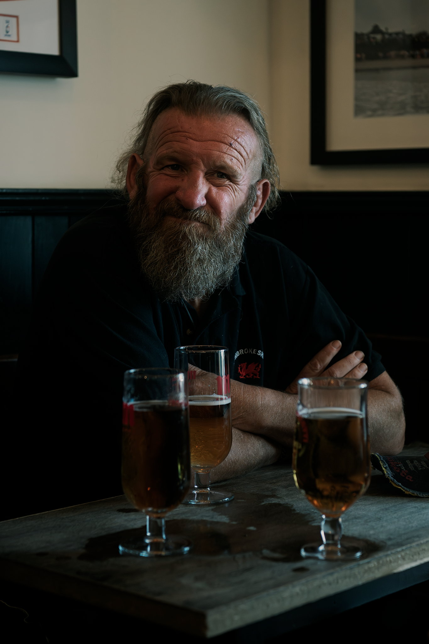 A bearded man with gray hair sitting at a table with three glasses of beer in front of him, smiling and crossing his arms inside a dimly lit pub or restaurant.