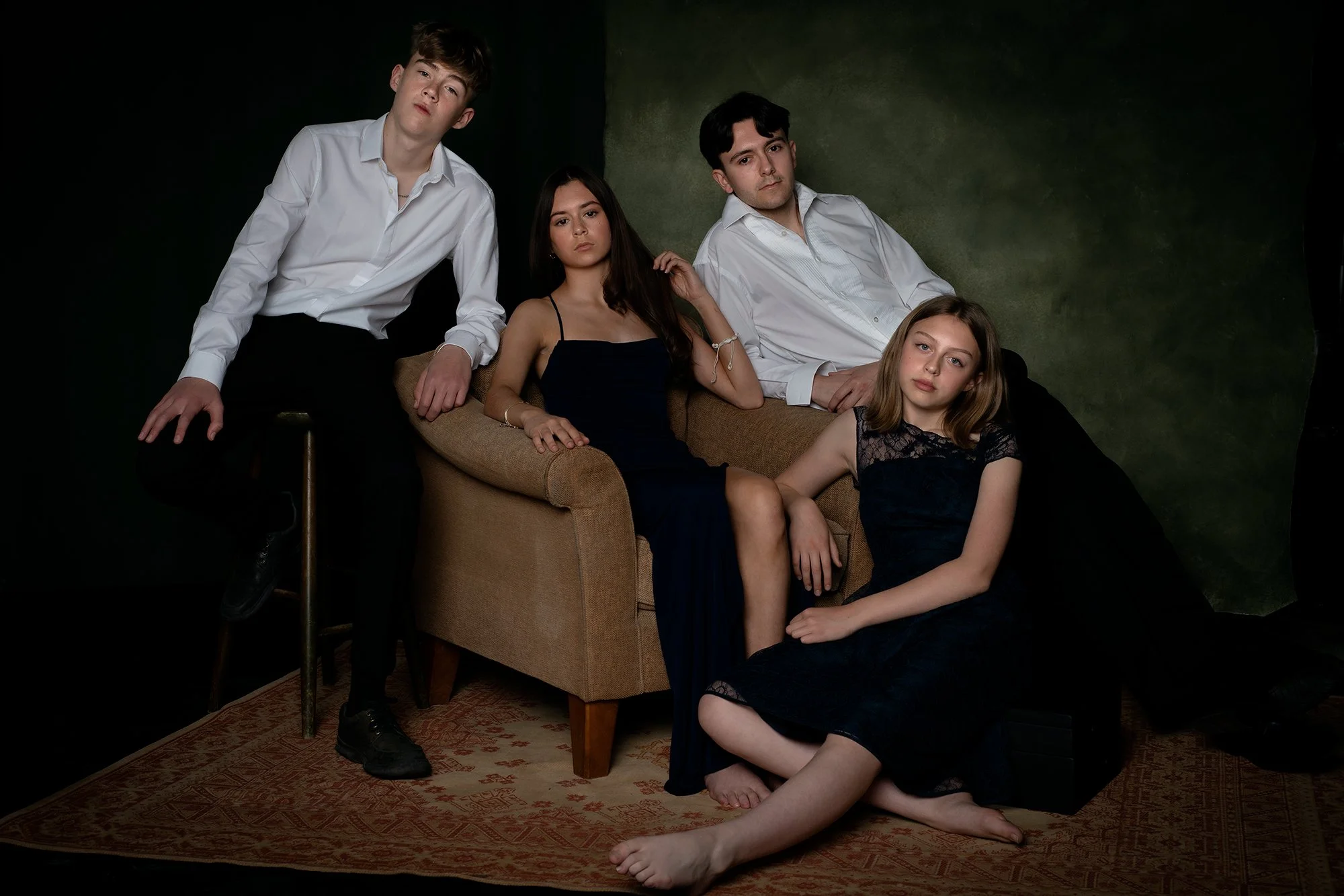 Group of four young people, three sitting and one leaning on a chair, in a dark room with a green and black backdrop, dressed in formal attire.