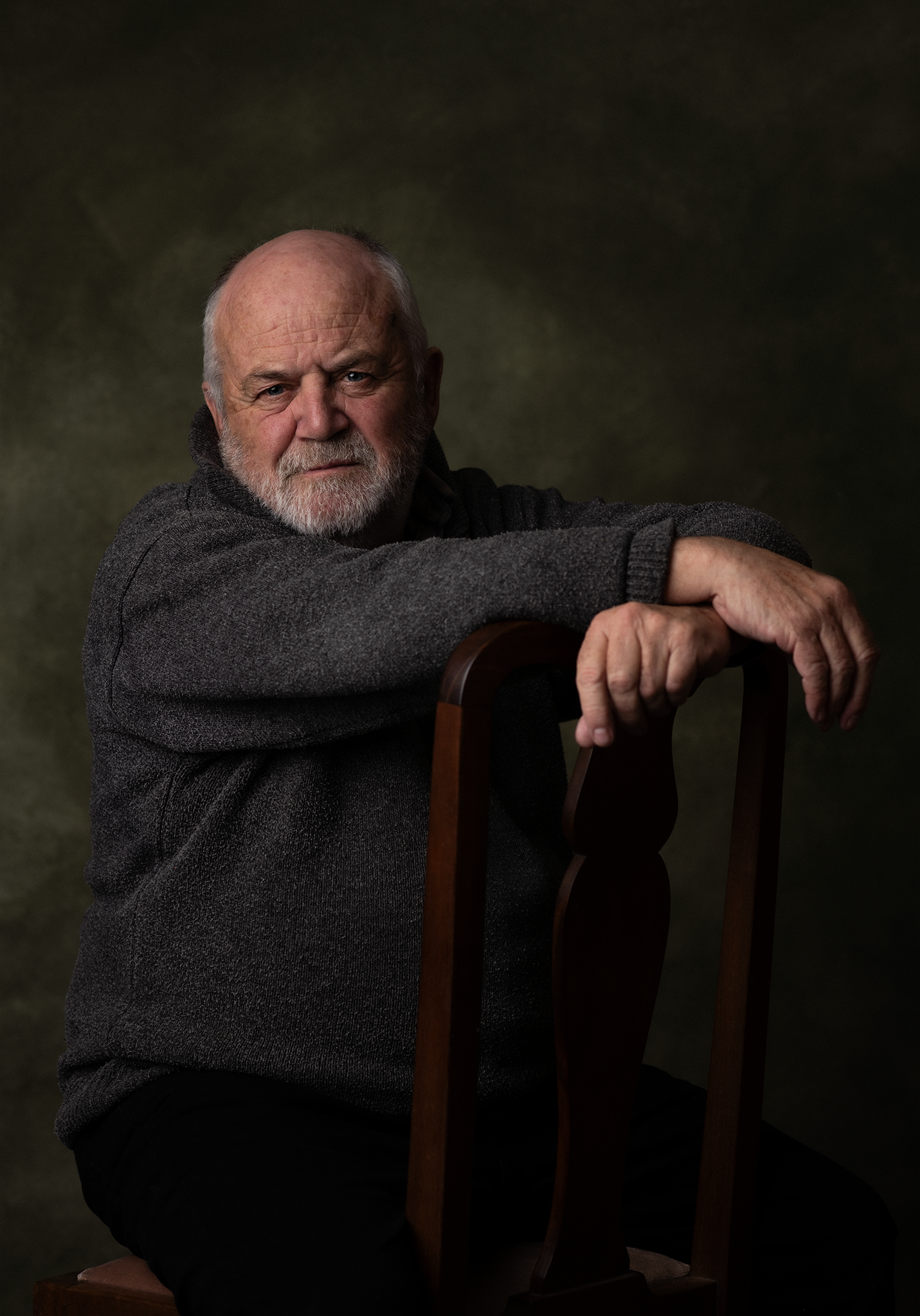 An older man with a white beard and receding hairline, wearing a dark gray sweater, sitting on a wooden chair with a dark green background. He is resting his arms on the top of the chair's backrest and looking directly at the camera with a serious ex