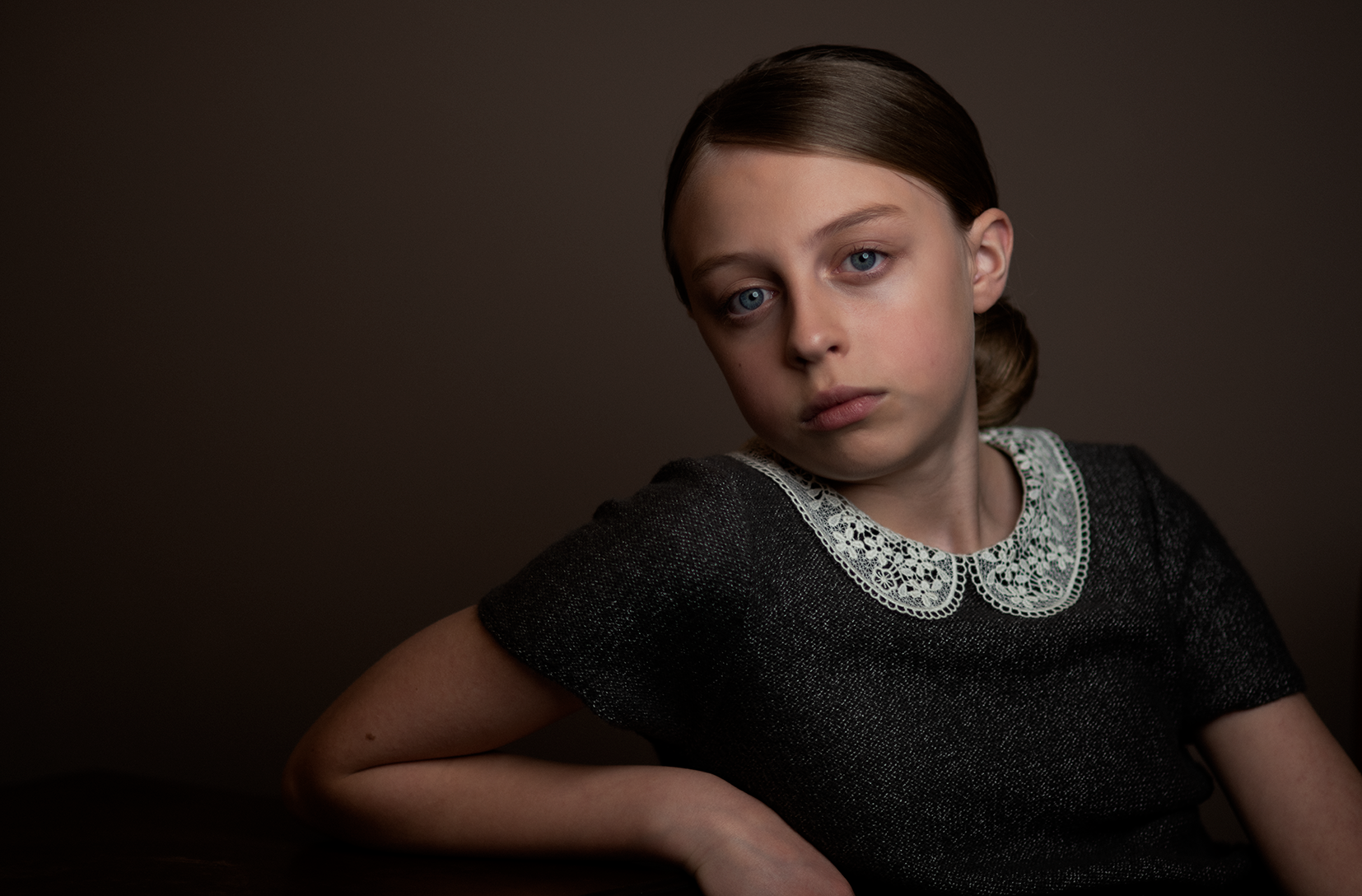 A young girl with light skin, blue eyes, and brown hair styled in a low bun, wearing a dark dress with a white lace collar, sits against a dark background, looking directly at the camera with a serious expression.