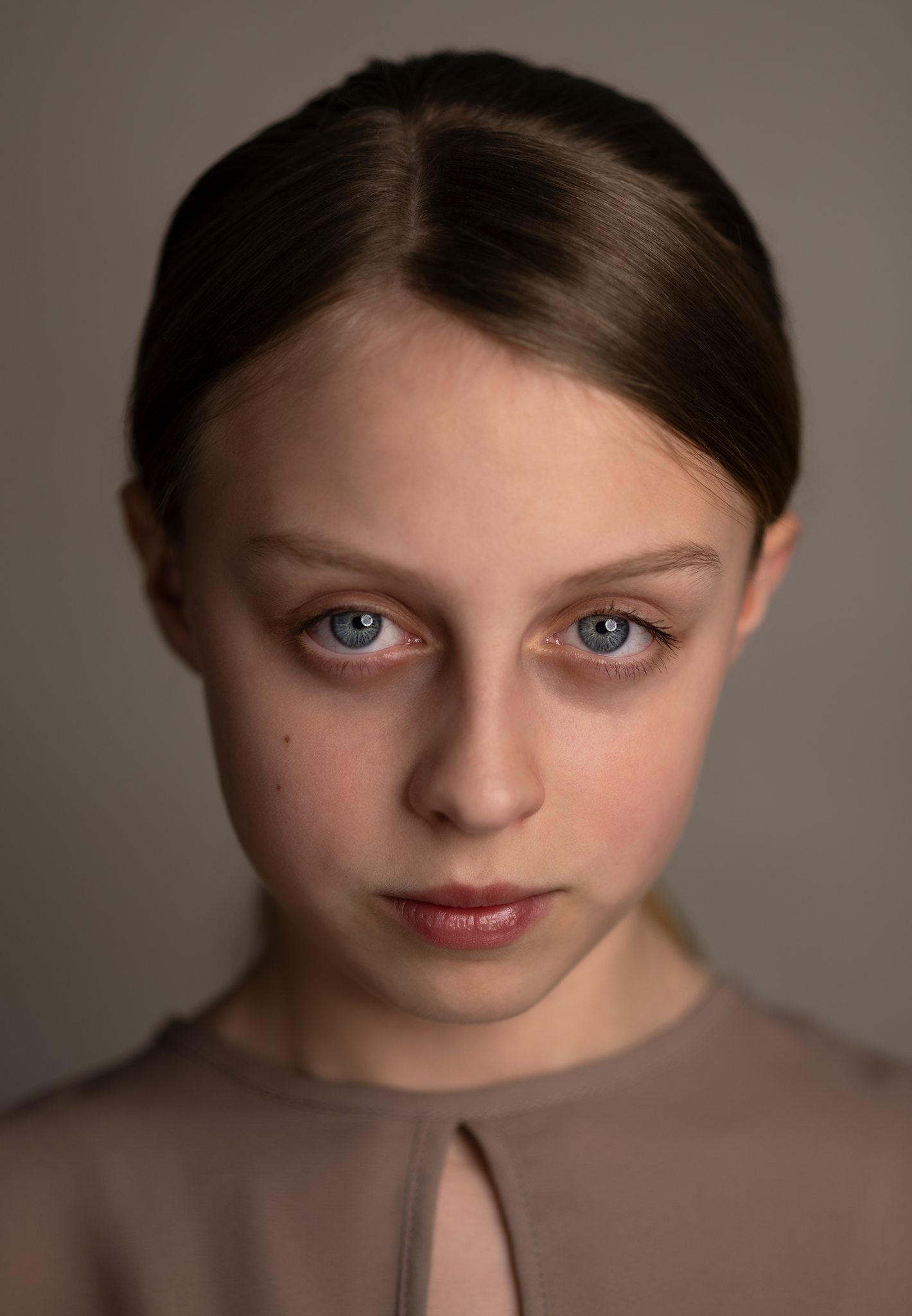 Close-up portrait of a young girl with blue eyes, light skin, and brown hair styled in a side part, wearing a taupe top, against a plain background.