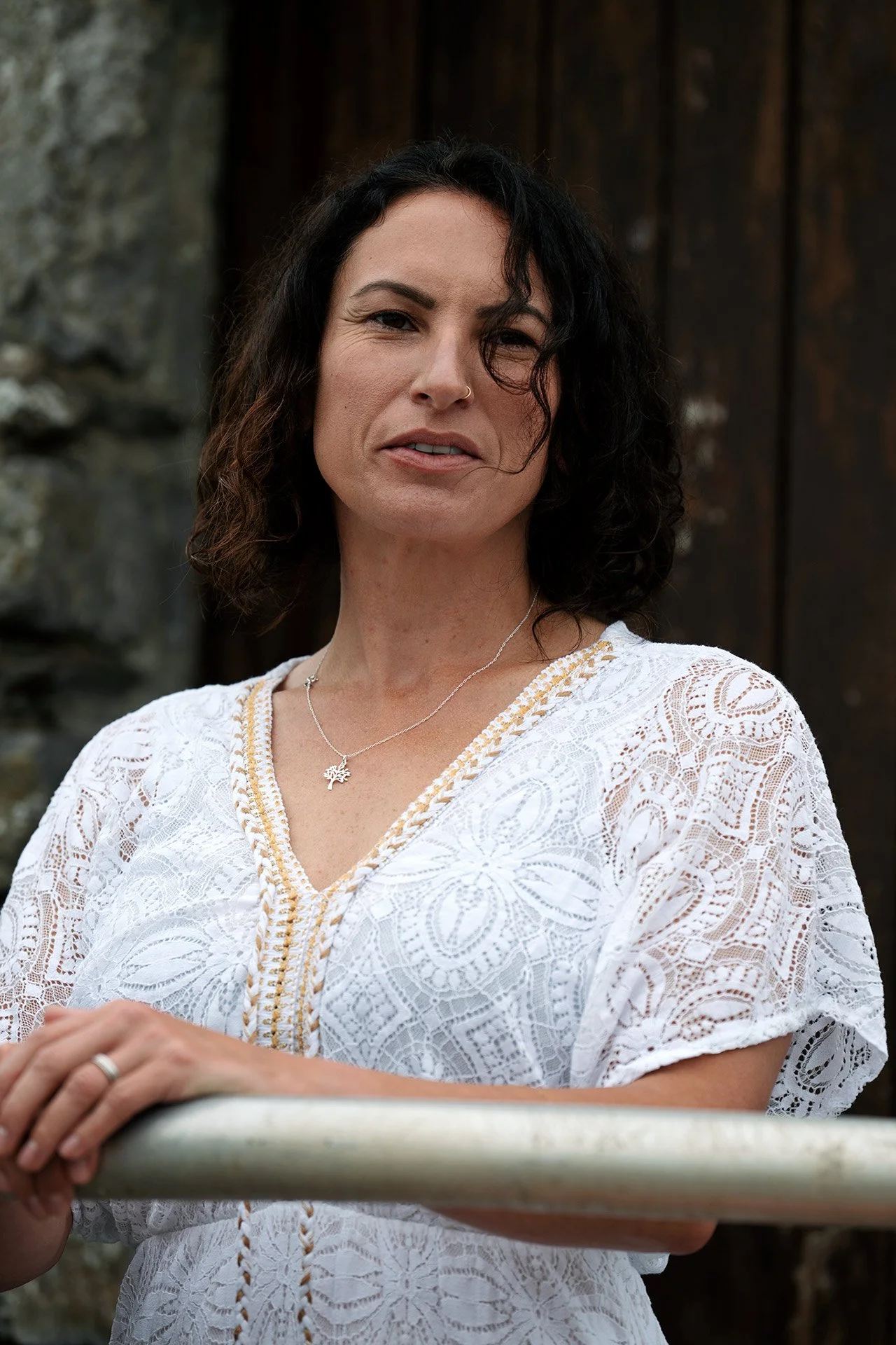 A woman with wavy dark hair, wearing a white lace dress with gold embroidery and a silver necklace with a tree pendant, standing outdoors near a metal railing with a stone wall in the background.