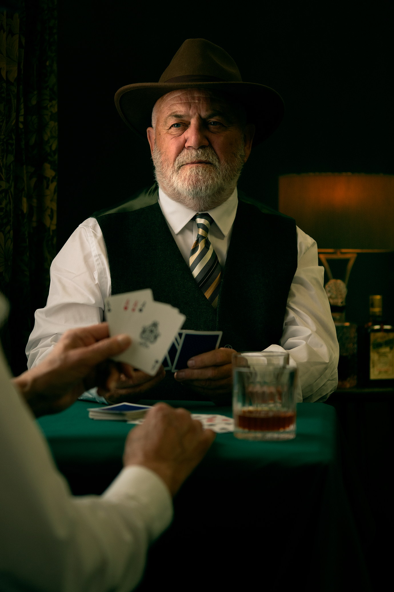 An elderly man with a white beard and mustache, wearing a brown fedora hat, white shirt, dark vest, and striped tie, sitting at a card table playing a card game with a glass of whiskey in front of him. Another player is showing hand of cards.