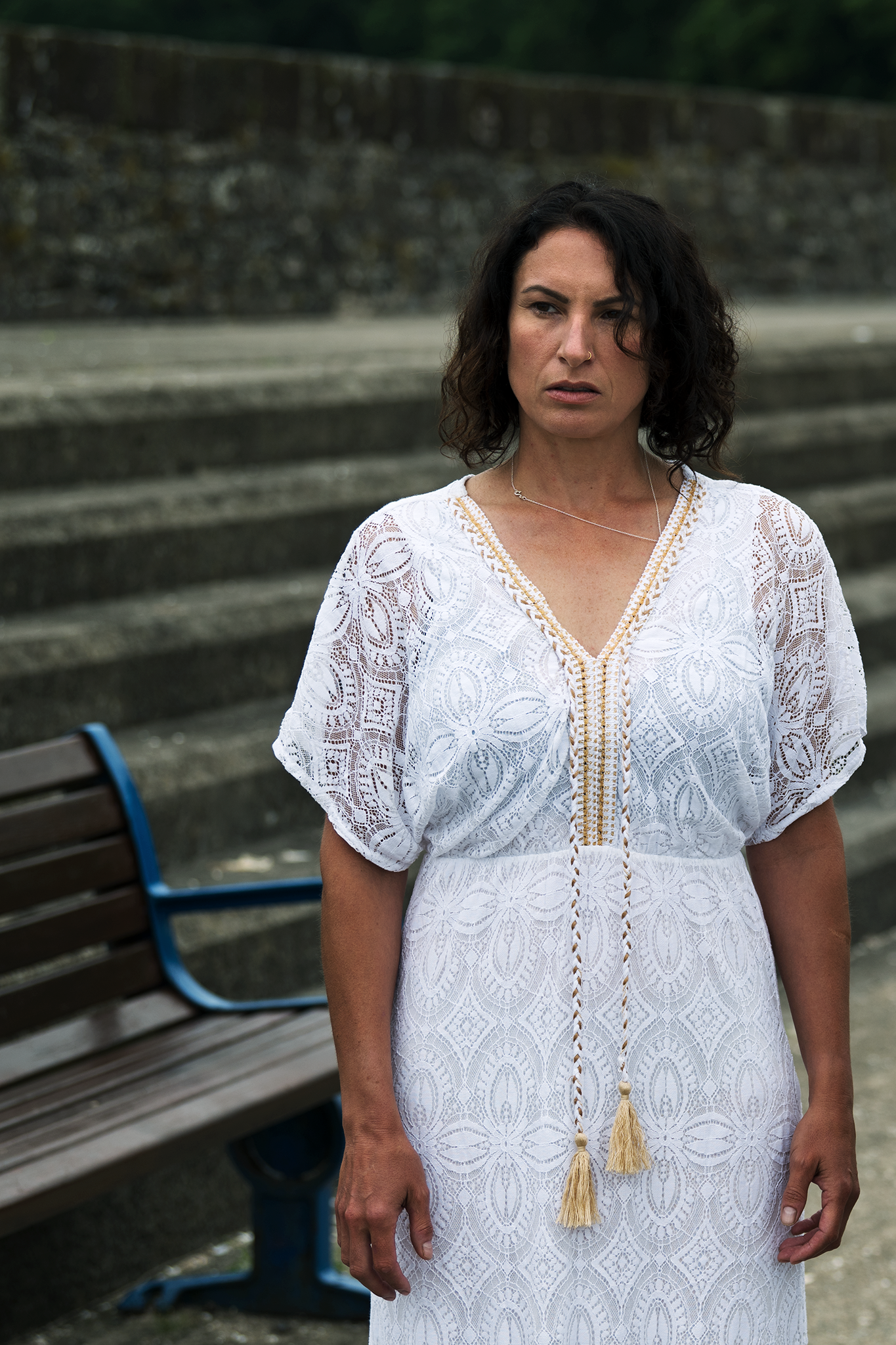 A woman with curly dark hair standing outdoors next to a bench, wearing a white lace dress and a gold-tassel necklace.