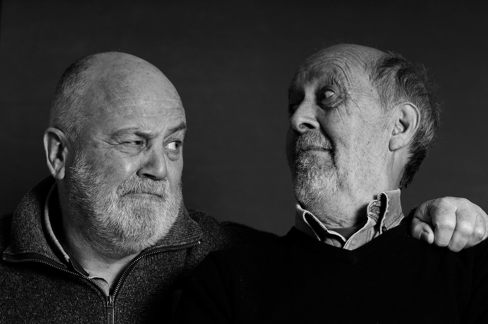 Two older men with beards, one with a bald head, the other with hair, looking at each other with one man's hand on the other's shoulder in a black and white portrait.