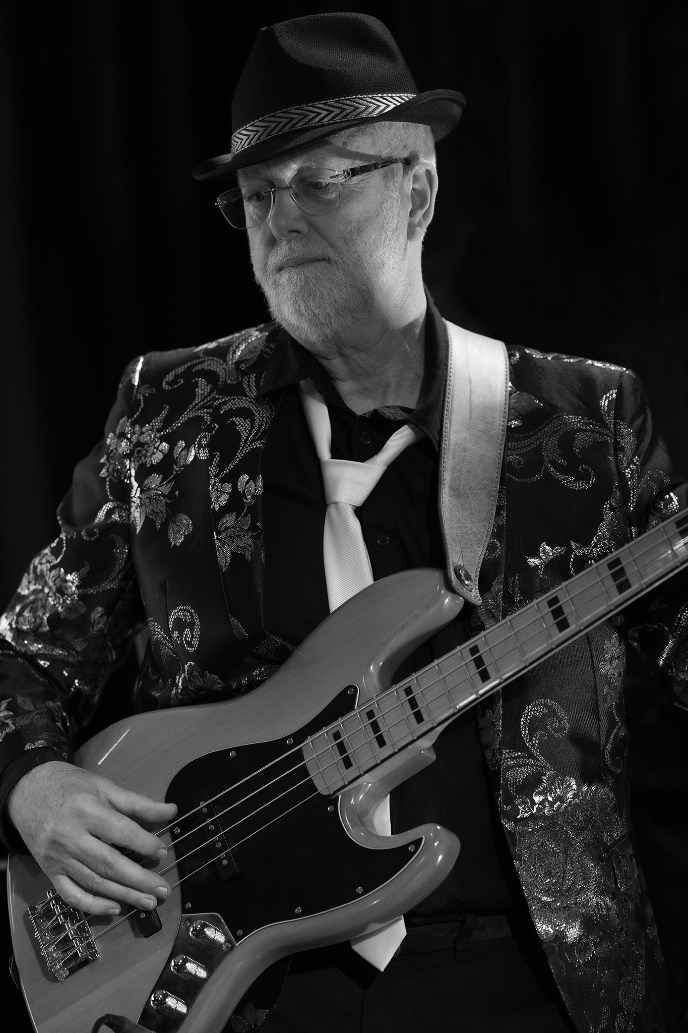 A senior man with a beard, wearing glasses, a patterned blazer, a hat with a chevron band, and a white tie, playing an electric bass guitar in a performance setting.