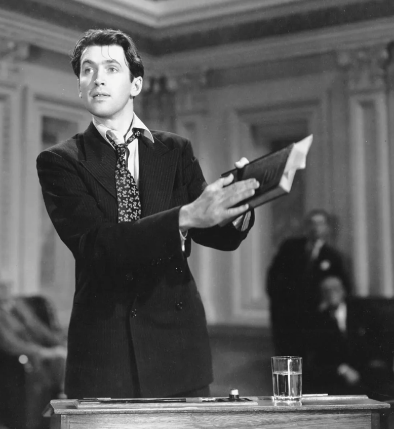 A black and white photograph of a young man in a suit holding a book, standing in what appears to be a courtroom or formal hearing room with ornate wood paneling, with a glass of water on the table in front of him.