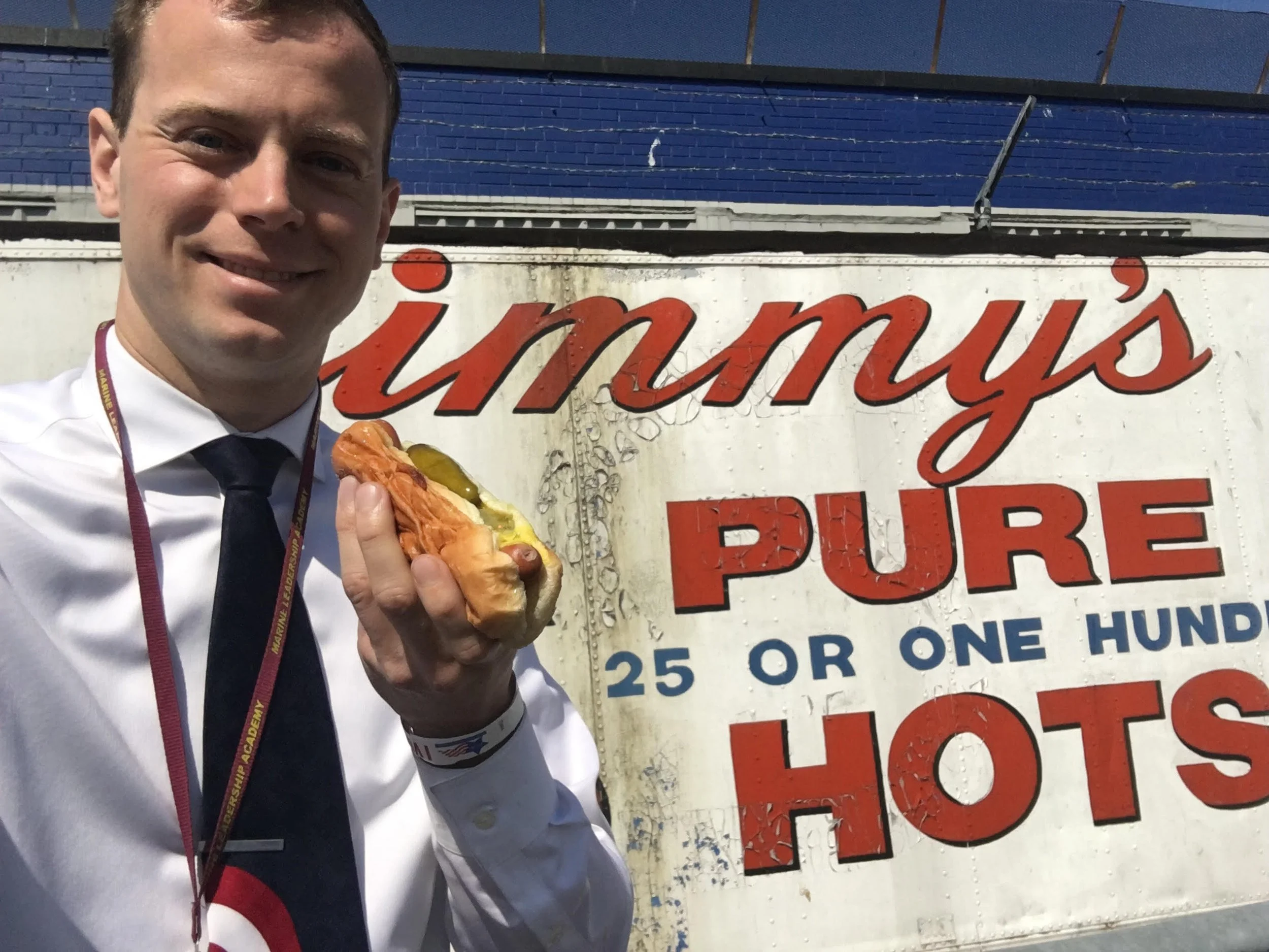 Man in white shirt and dark tie smiling and holding hot dog with mustard in front of a sign that reads 'Jimmy's Pure 25 or One Hundred Hot.'