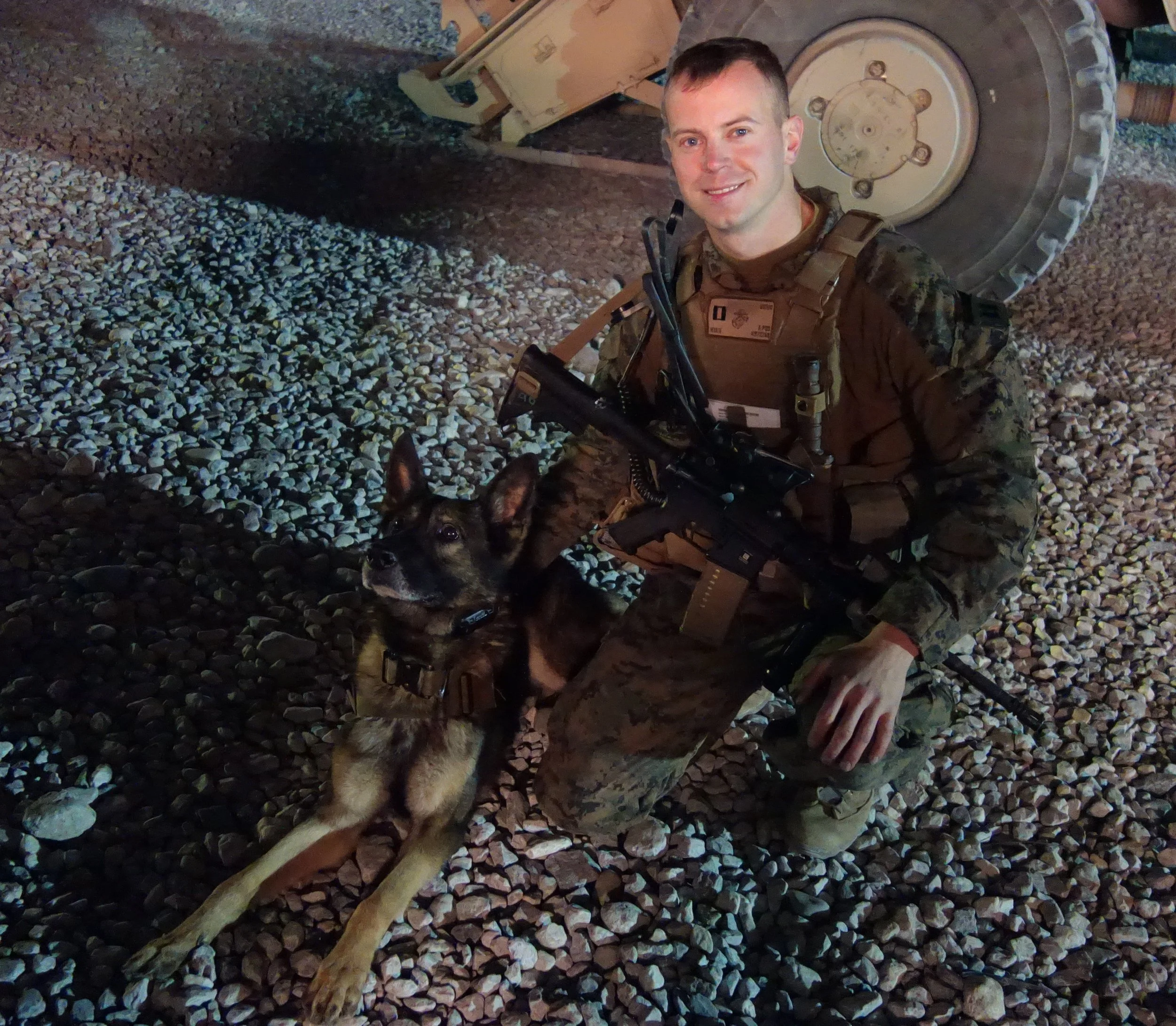 A soldier in camouflage uniform kneels on gravel ground next to a military working dog. The soldier holds a rifle and wears tactical gear, smiling at the camera. A large military vehicle is in the background.