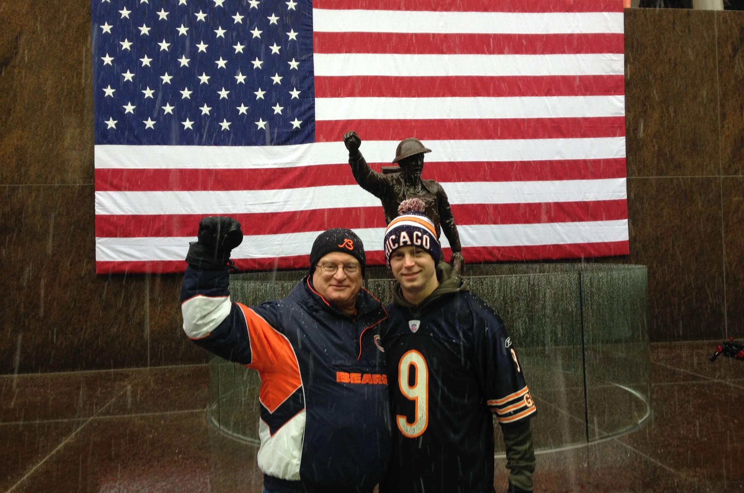 Two men standing in front of the American flag at the National World War II Memorial, dressed in Chicago Bears gear during rainy weather.