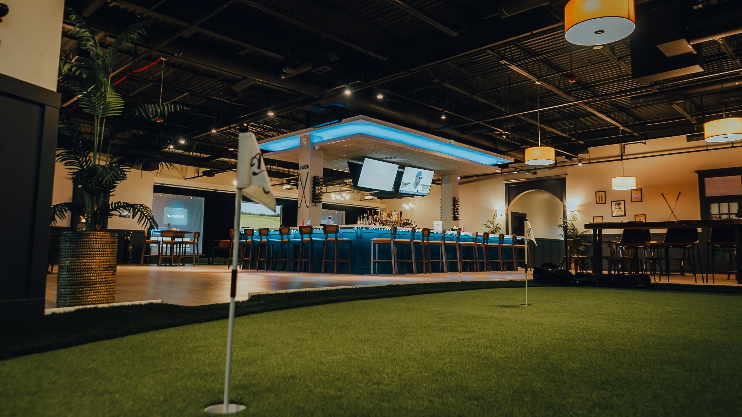 Indoor golf simulation venue with a putting green in the foreground and a bar area with high chairs and televisions in the background.