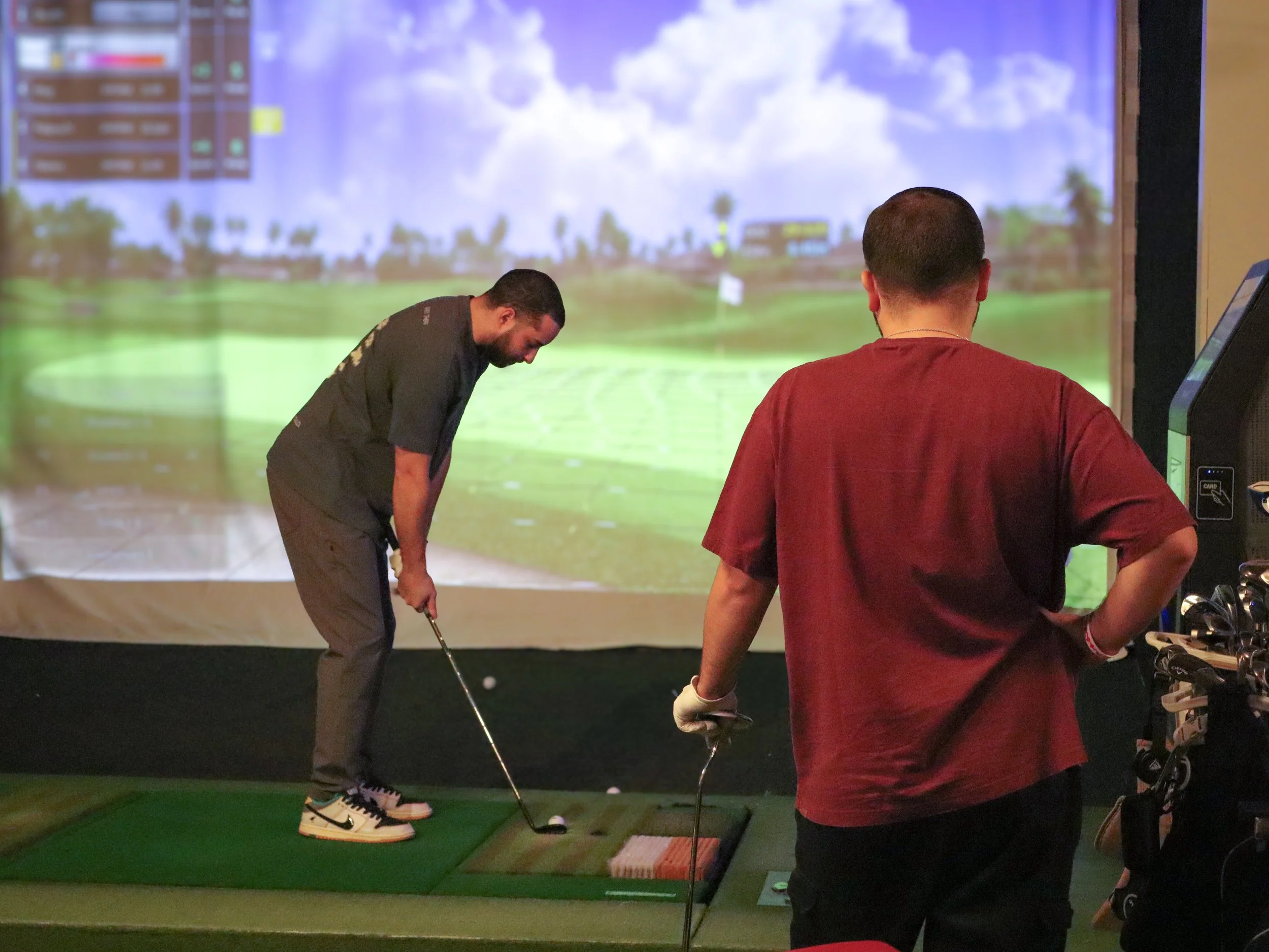 Two men at a golf simulator. One man is swinging a golf club, while the others are talking or observing.