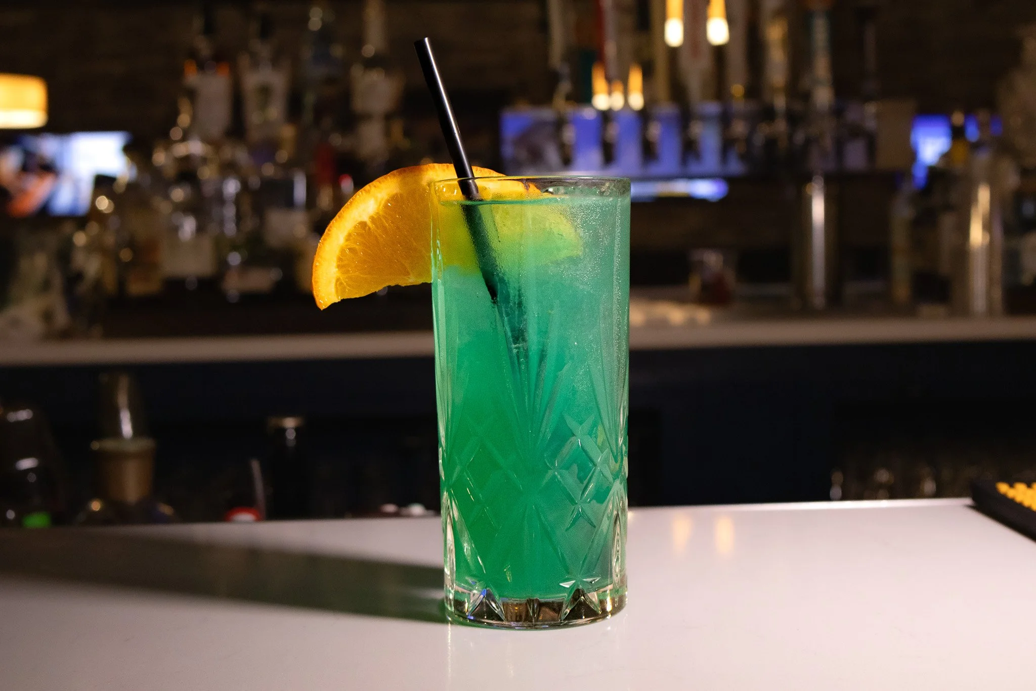 A tall glass cocktail with greenish-blue color, garnished with an orange wedge and a black straw, set on a bar counter with a blurred bar background.