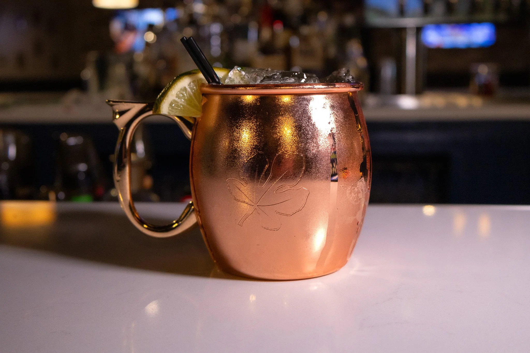 A copper mug with a lime wedge and black straw, filled with a cold drink, placed on a bar counter. The mug has a leaf engraving on it and condensation on the outside.