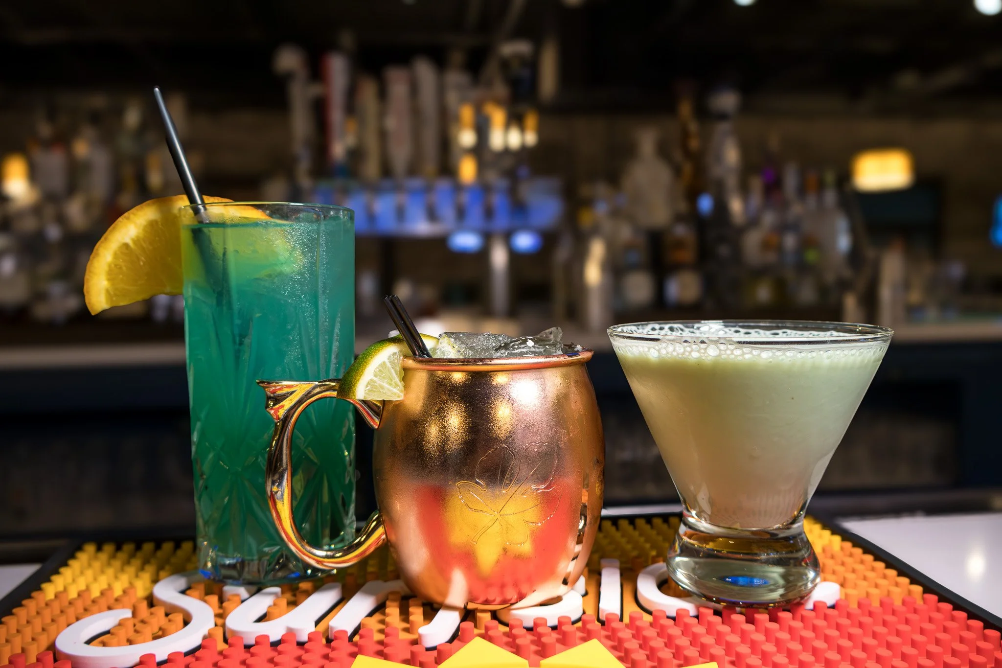 Three cocktails on a bar counter with a blurred bar background: a tall blue drink with an orange wedge, a copper mug with a lime wedge, and a pale green drink in a martini glass.