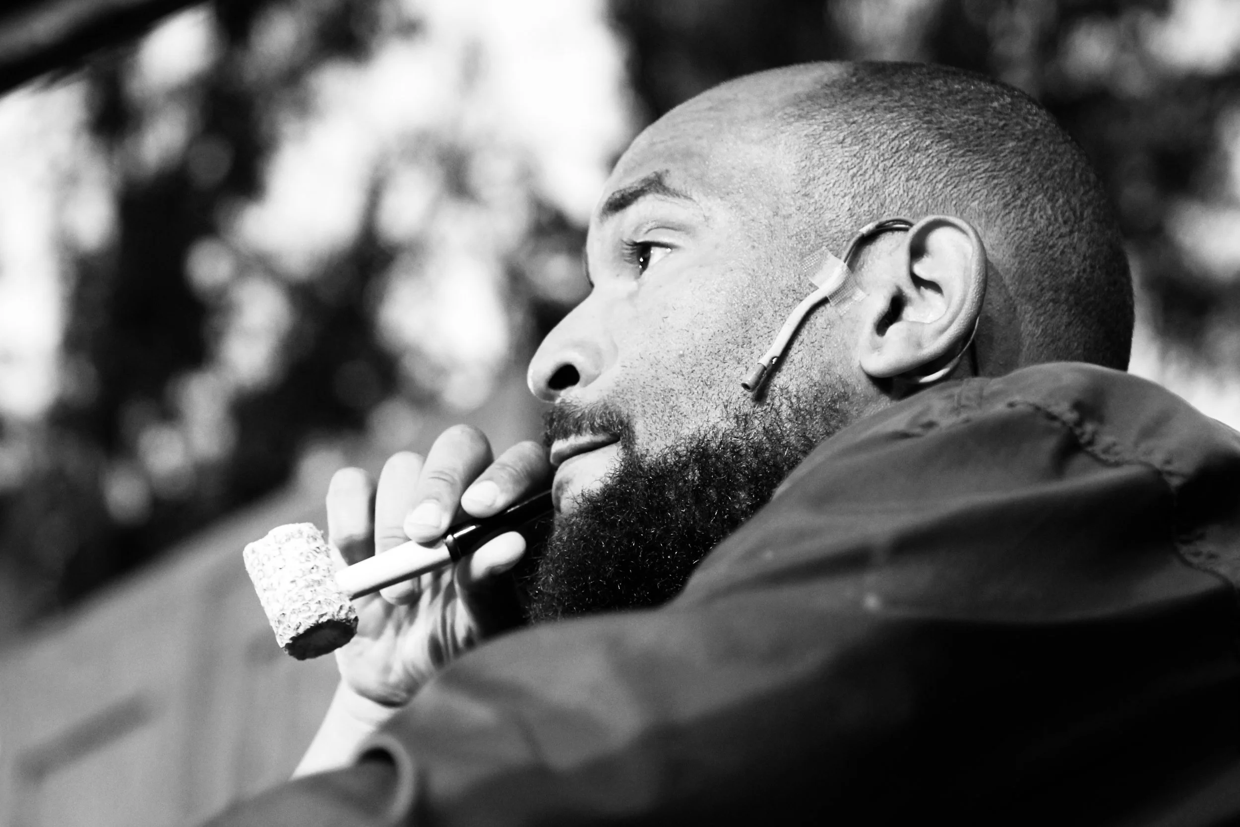A man with a beard and a close-cropped haircut is holding a smoking pipe near his lips. He has a hearing aid in his right ear and is wearing a jacket. The background is blurred.