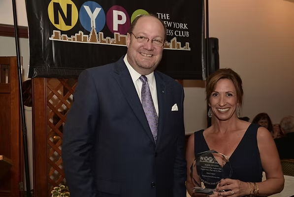 A man and woman dressed formally, smiling at the camera, at an NYPD event, with an NYPD banner in the background.