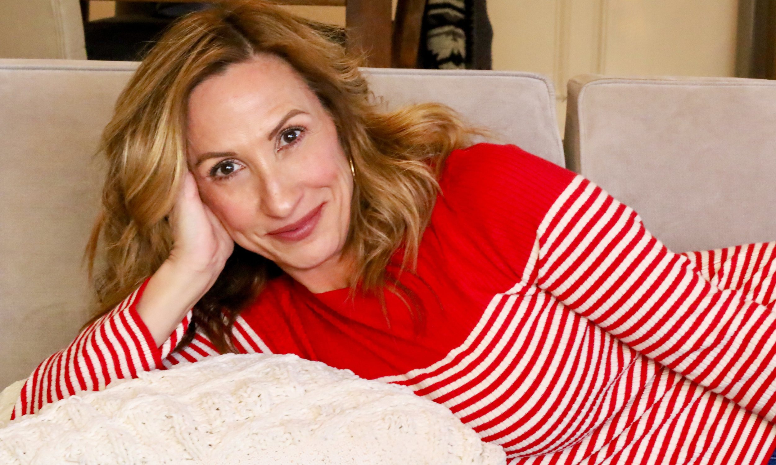 A woman with wavy red hair lying on a beige couch, wearing a red and white striped long sleeve shirt, resting her head on her hand and smiling at the camera.