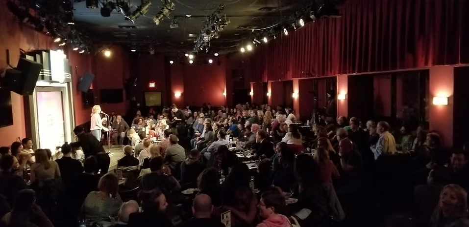 A comedy club with a large audience seated at tables, facing a stage where a performer is speaking into a microphone.