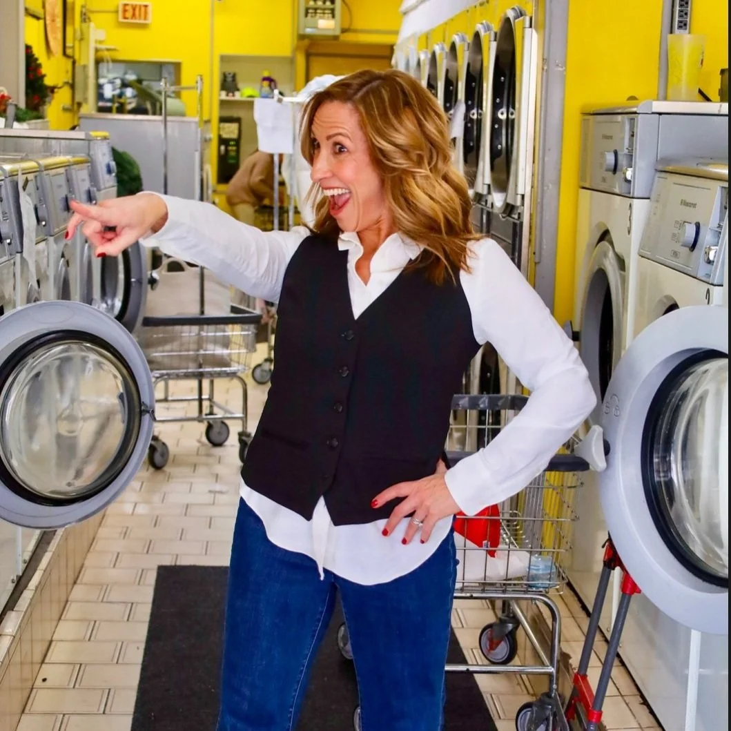 A woman with shoulder-length wavy hair wearing a white shirt and black vest, happily pointing at a washing machine in a laundromat with yellow walls.