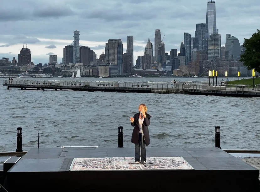 Becky Veduccio on stage by the water with New York City skyline in the background.