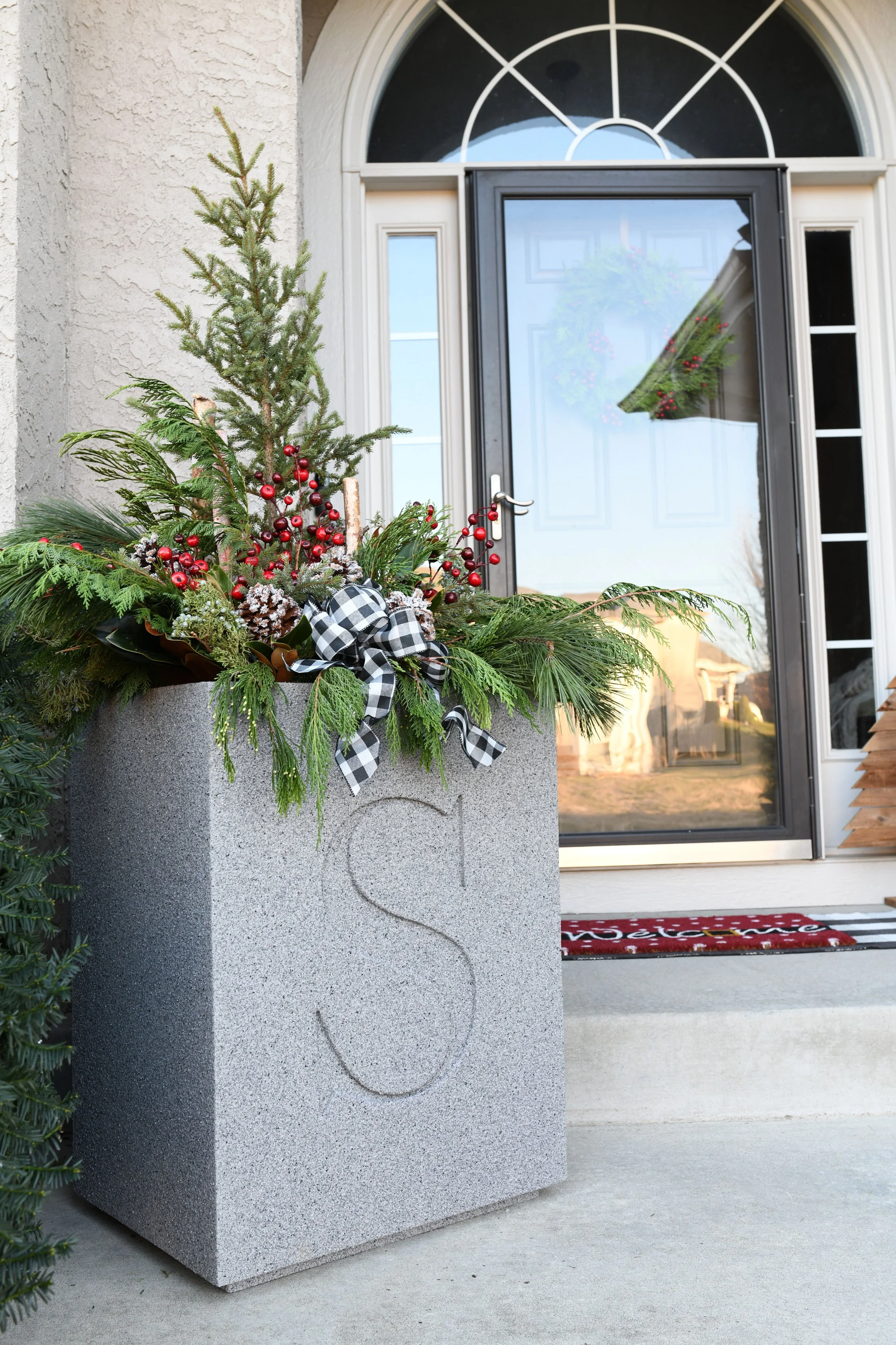 Decorative holiday planter with pine branches, red berries, pinecones, and a black-and-white checked bow in front of a house with a black door and Christmas wreath.