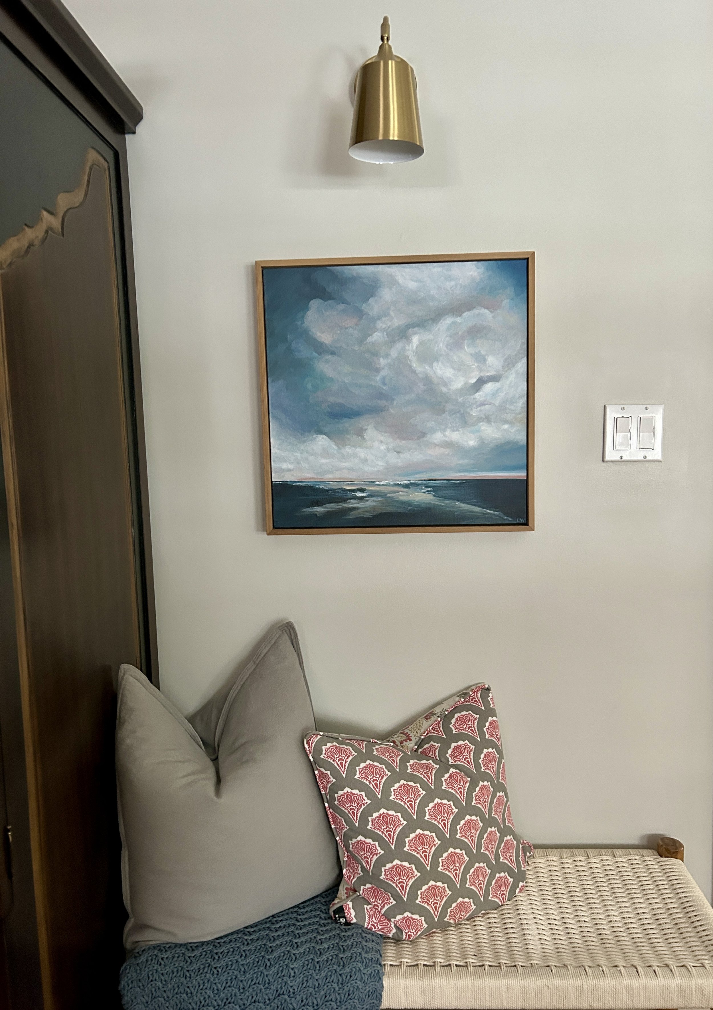 Interior corner with a framed ocean painting, brass wall lamp, woven bench, two decorative pillows, and a textured blanket.