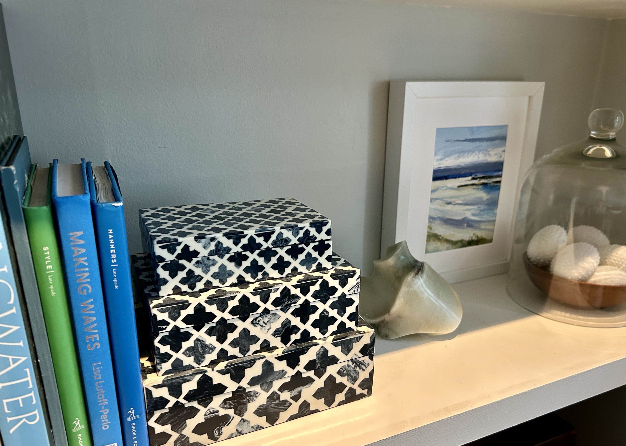 Books and decorative items on a shelf, including patterned boxes, a conch shell, framed picture, and a glass dome with white objects.