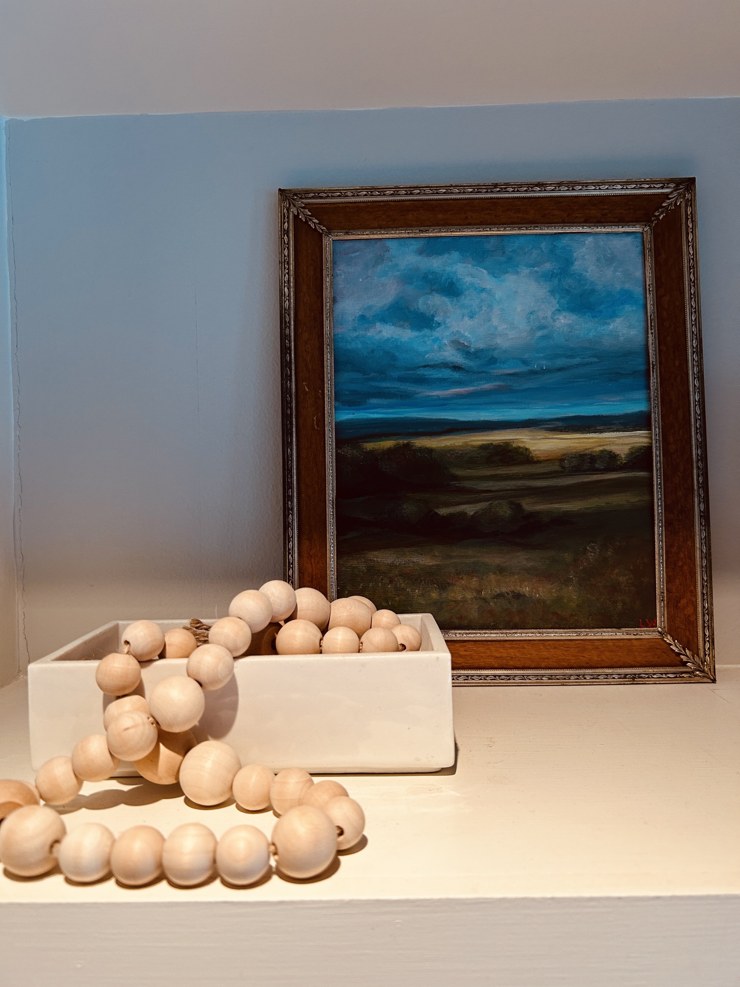 A framed painting of a landscape with a sky on a shelf, next to a white rectangular container holding a string of wooden beads.