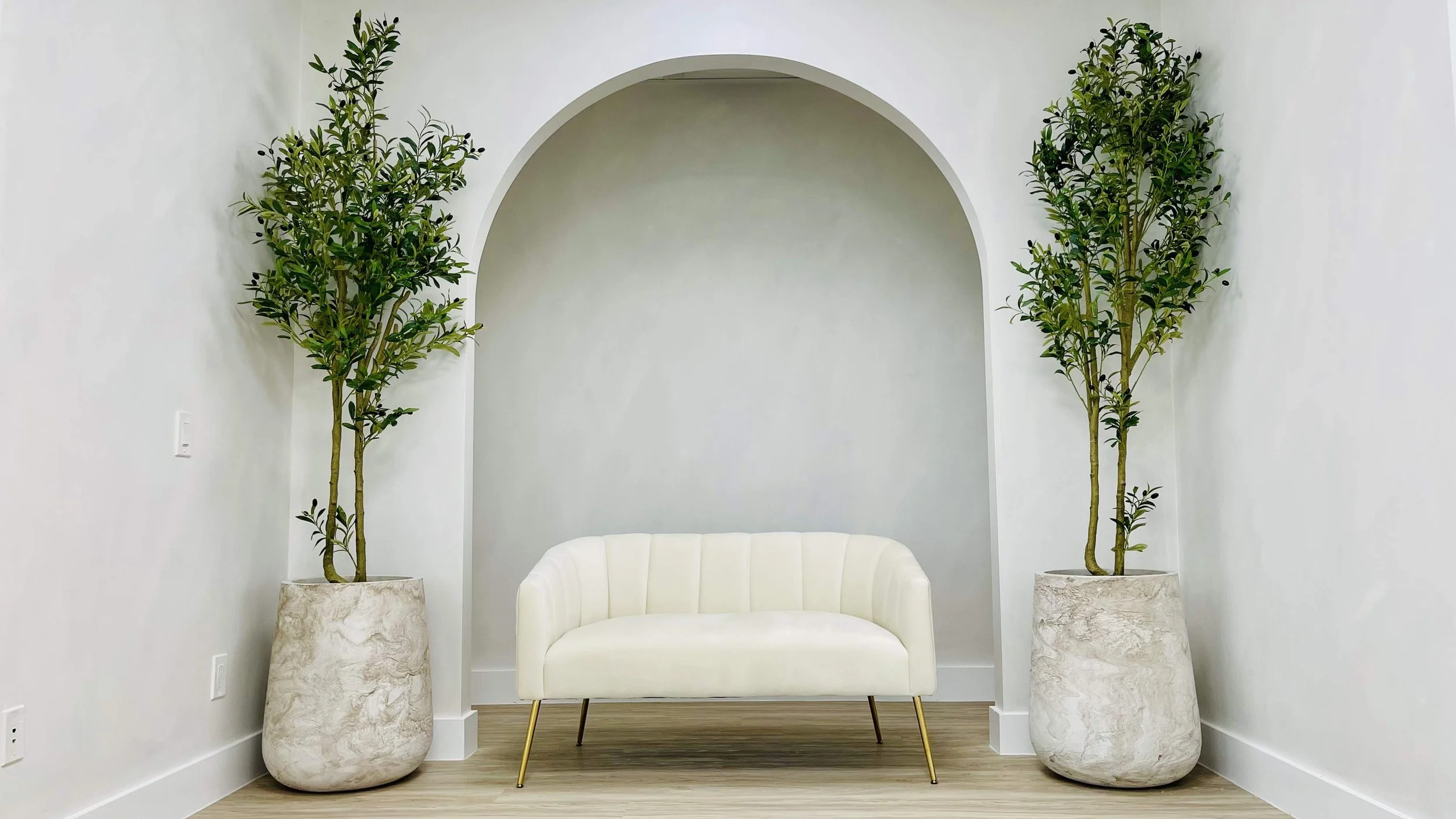 White modern curved sofa between two tall potted green plants against a white wall with an arched alcove.