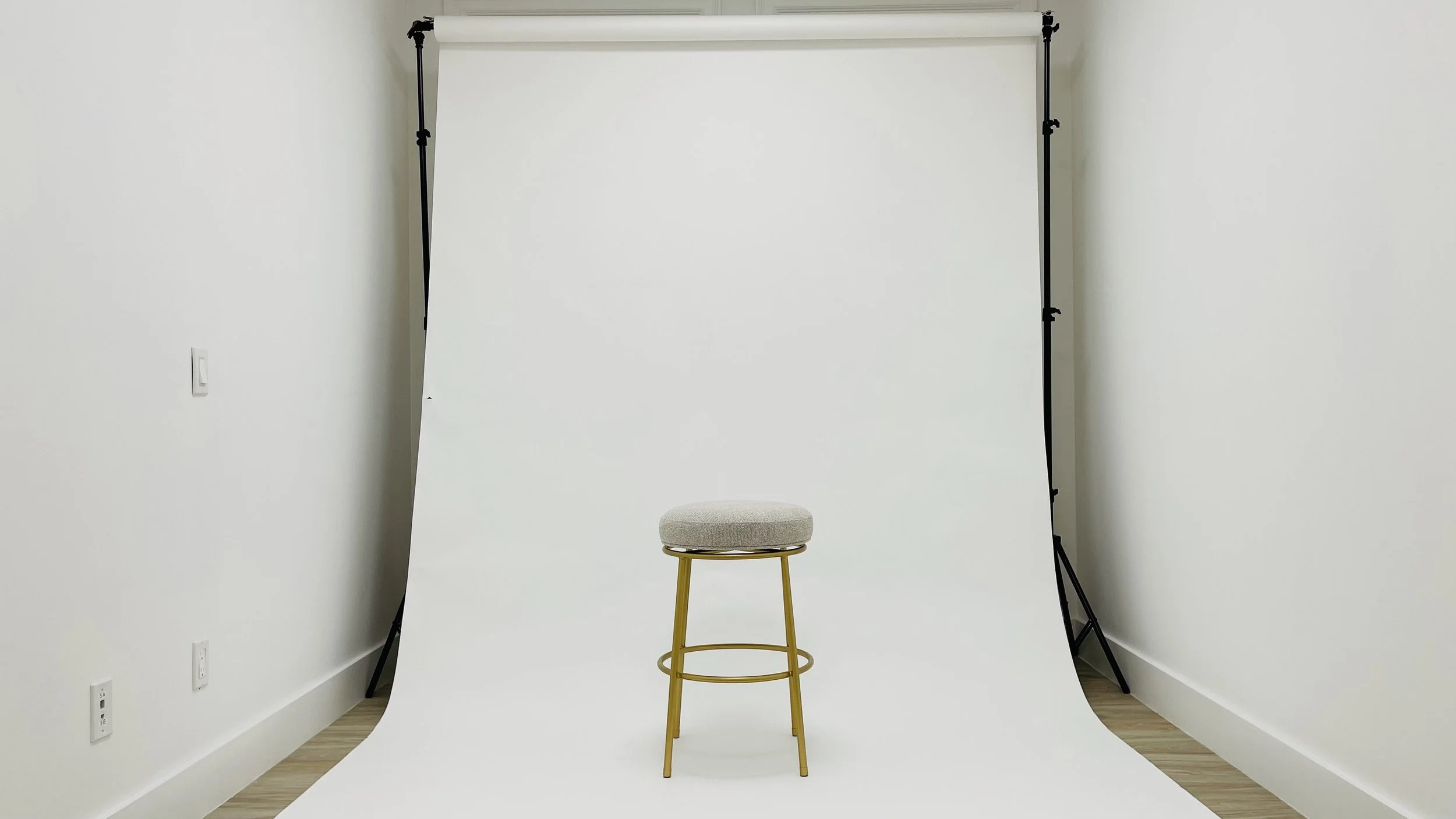 A photography studio with a white backdrop, a beige cushioned stool with gold legs positioned in front of the backdrop.