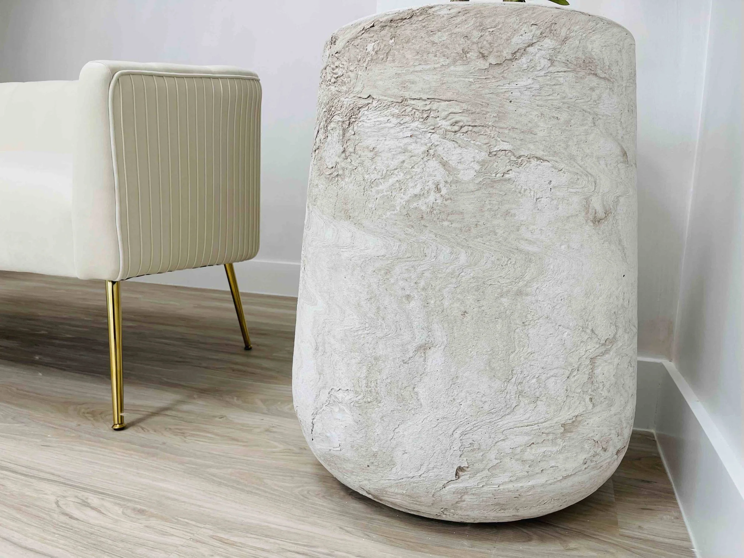 A large, rounded beige stone sculpture placed on a wooden floor next to a white and beige upholstered chair with gold metal legs.