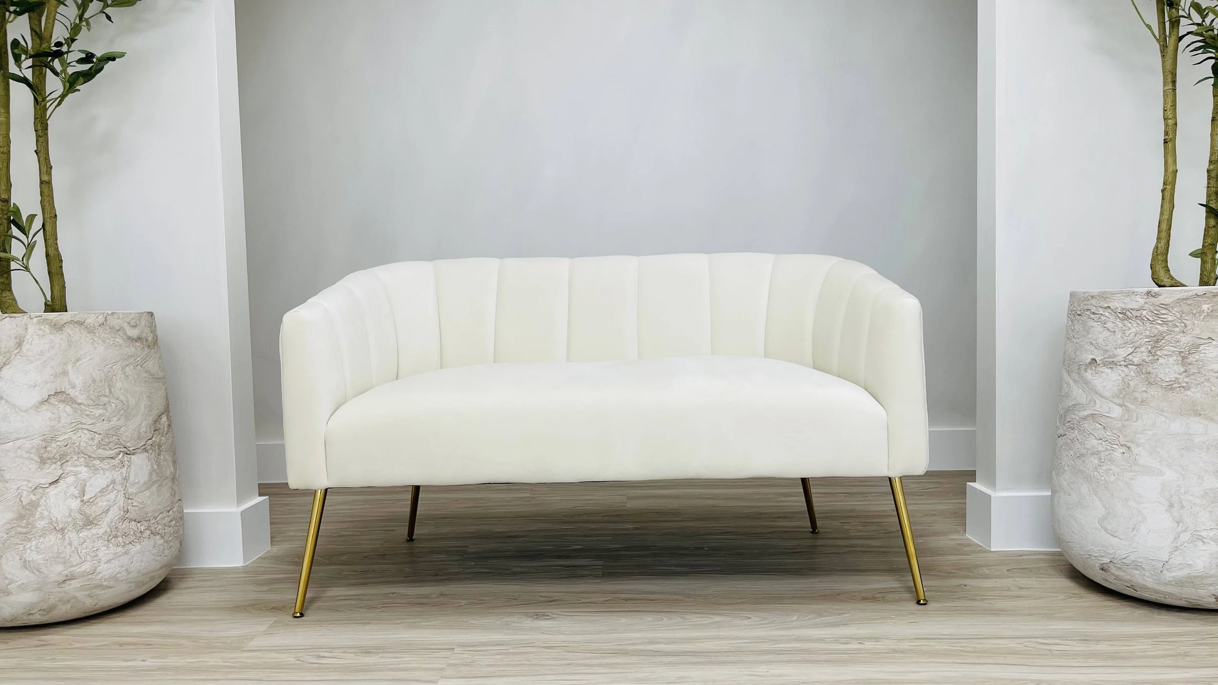 White modern sofa with four gold and black legs, flanked by large, textured white planters with green plants, against a minimalist light gray wall and light wood flooring.