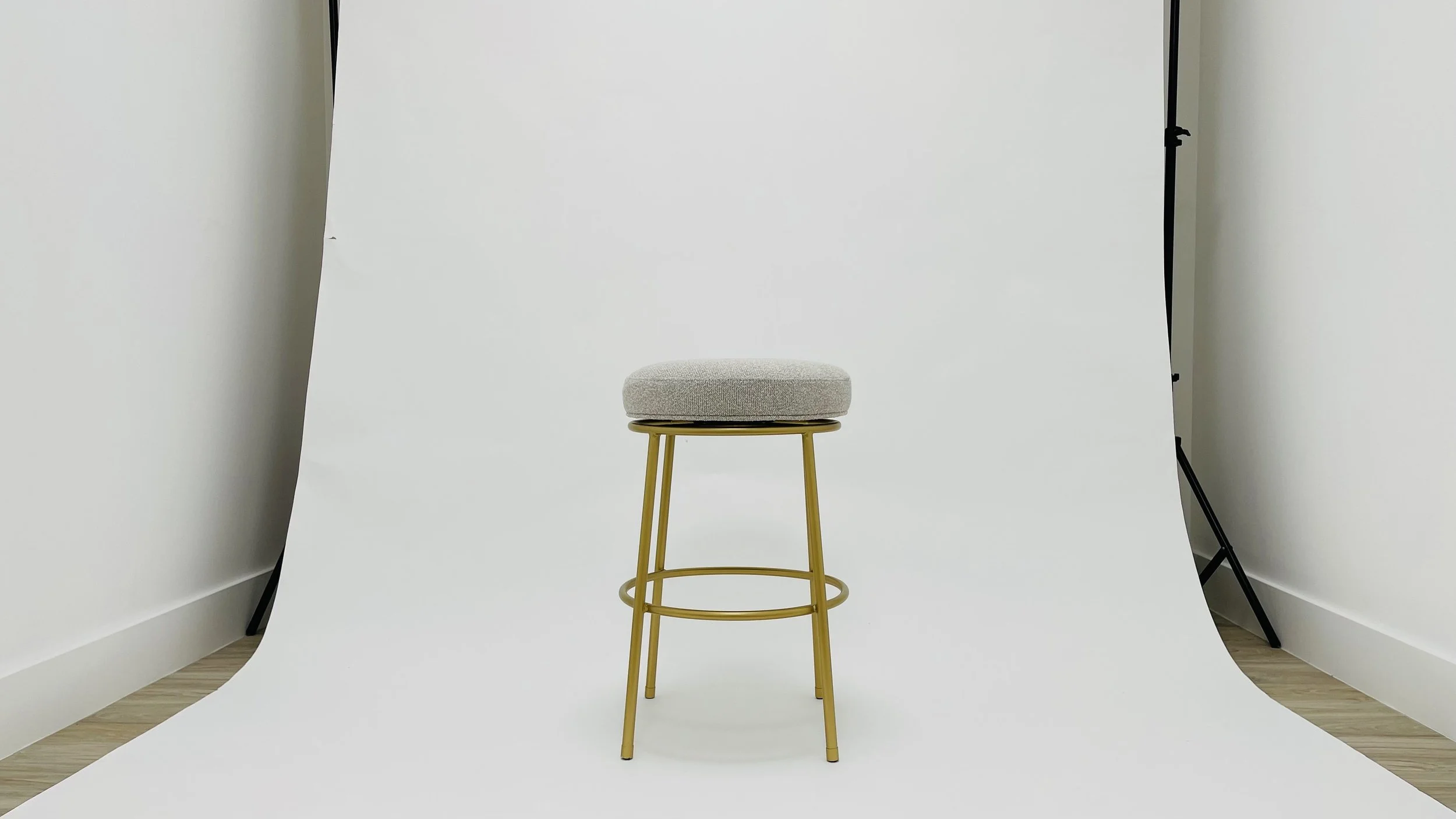 A beige cushioned stool with a gold metal frame and circular footrest, centered in front of a white backdrop in a photo studio.