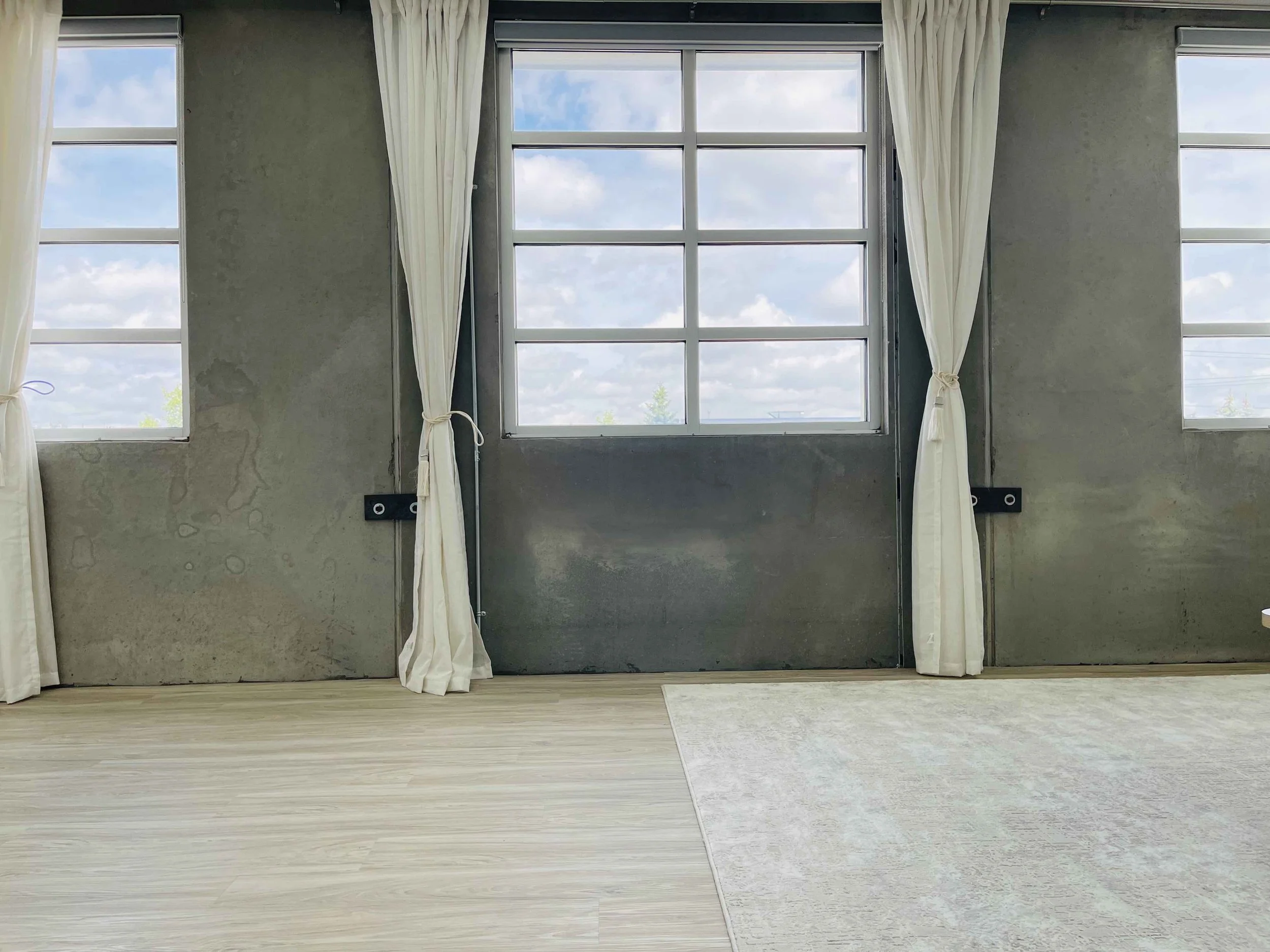 Interior room with large windows and cream curtains, concrete walls, light wood floor, and part of a light-colored rug.