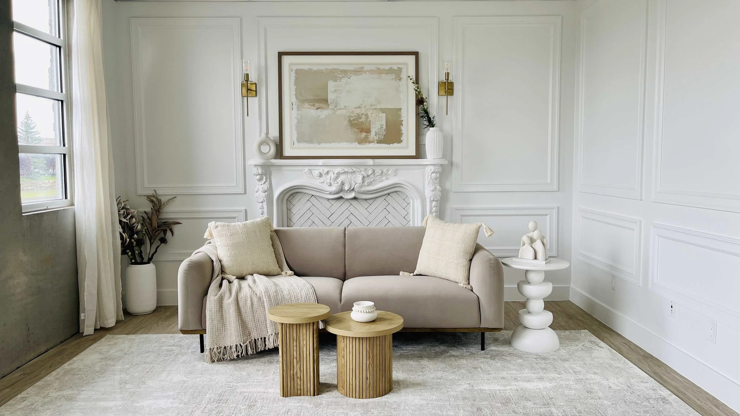 Modern living room with beige sofa, two wooden side tables, white decorative vase, framed abstract art above fireplace, and large window with cream curtains.