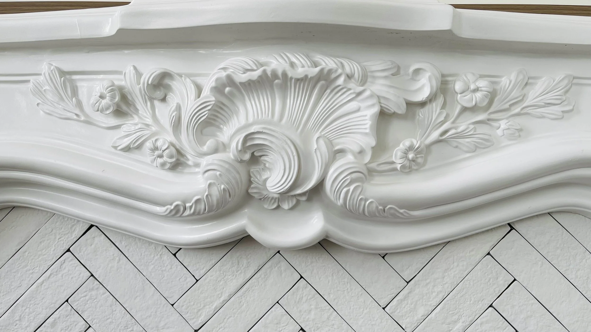 Close-up of ornate white decorative molding with floral and shell motifs above white tiled herringbone-patterned wall.