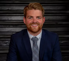 Portrait of a smiling man with a beard wearing a gray suit jacket, outdoors in an urban setting with modern buildings in the background.