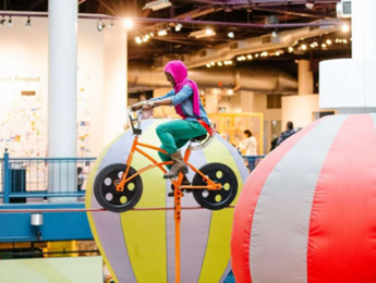 A person wearing a pink hijab and teal clothing is riding a bicycle-shaped ride at an indoor amusement park.