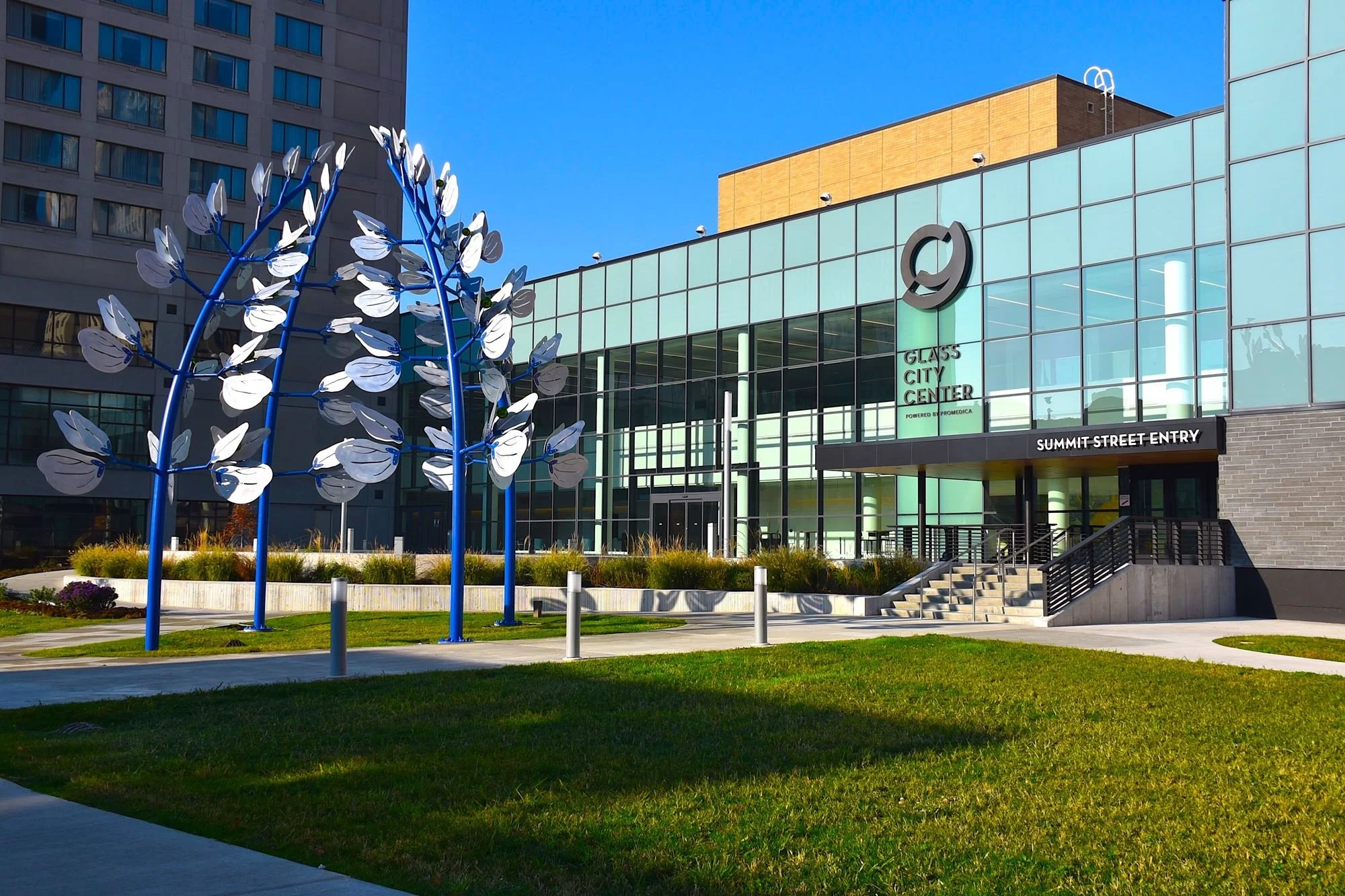 Exterior view of Glass City Center with modern glass facade, colorful outdoor sculpture resembling blue tree with white leaves, green landscaping, and clear blue sky.