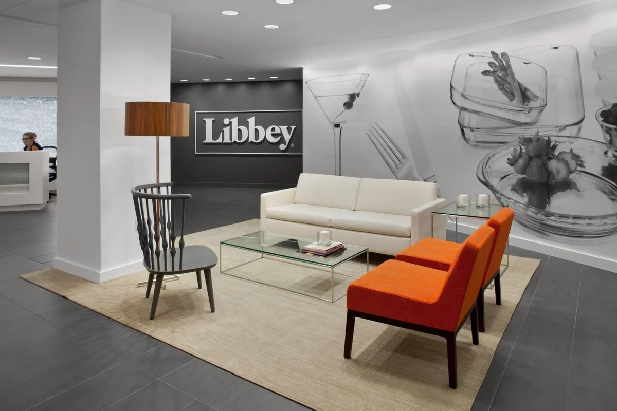 Modern office lobby with white sofa, orange armchairs, a wooden floor lamp, a glass coffee table, a black and white "Libbey" sign on a wall, and large black-and-white food-related artwork behind.