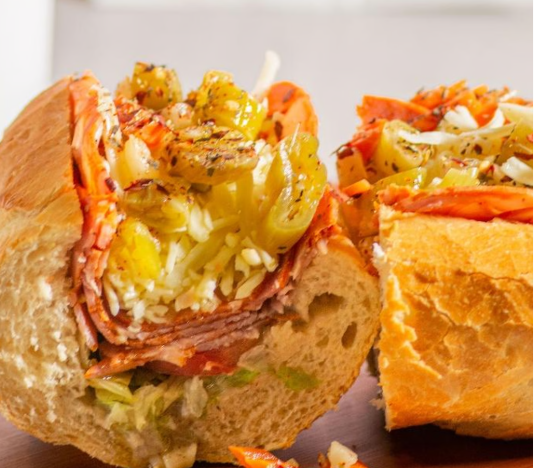 A sandwich with sliced meats, shredded cheese, pickles, and lettuce inside a toasted sub bun, next to a piece of cheesy bread with shredded cheese and vegetables.