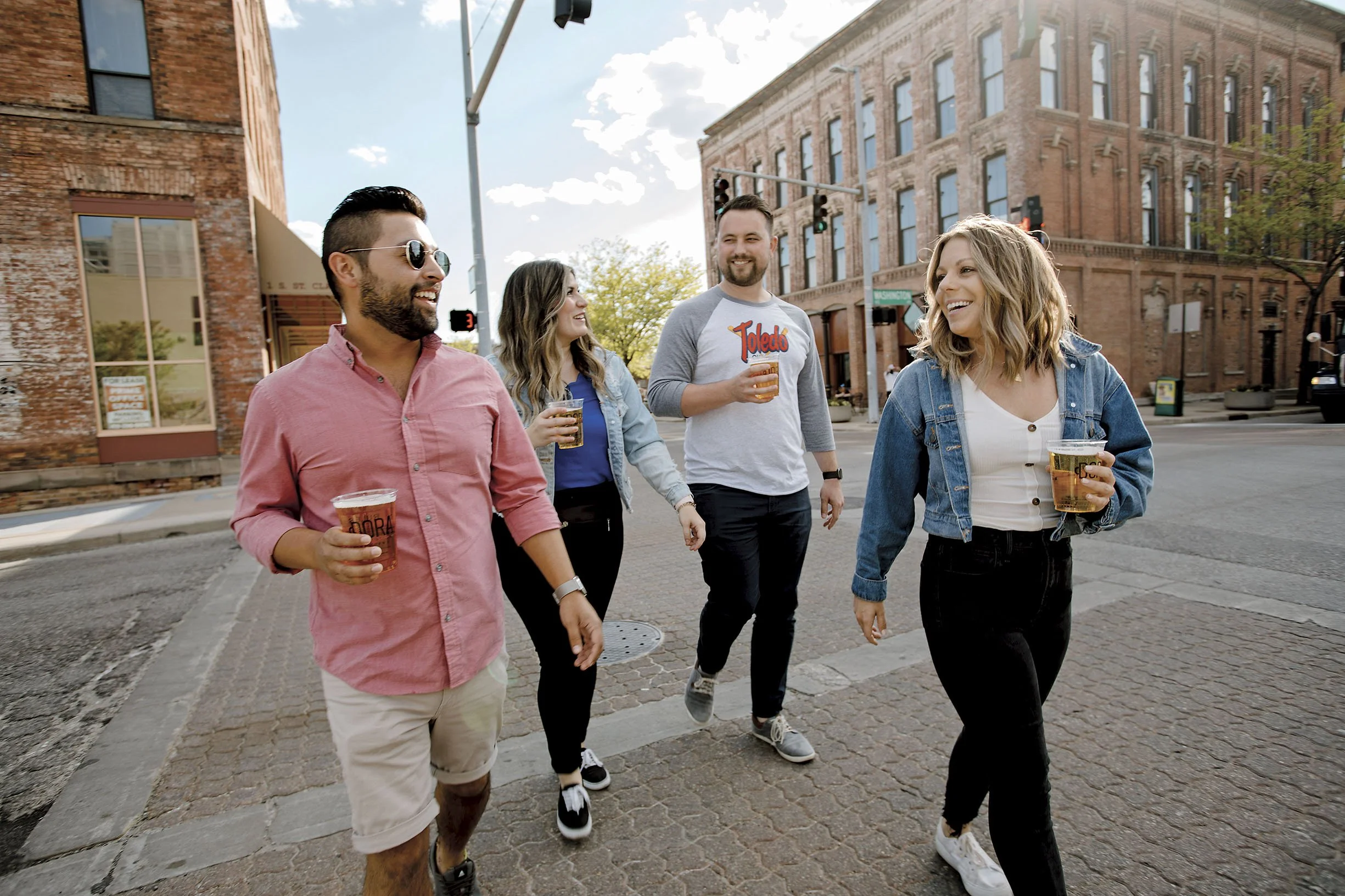 Four friends walking on a city street, smiling, and holding drinks in plastic cups, with old brick buildings in the background on a sunny day.