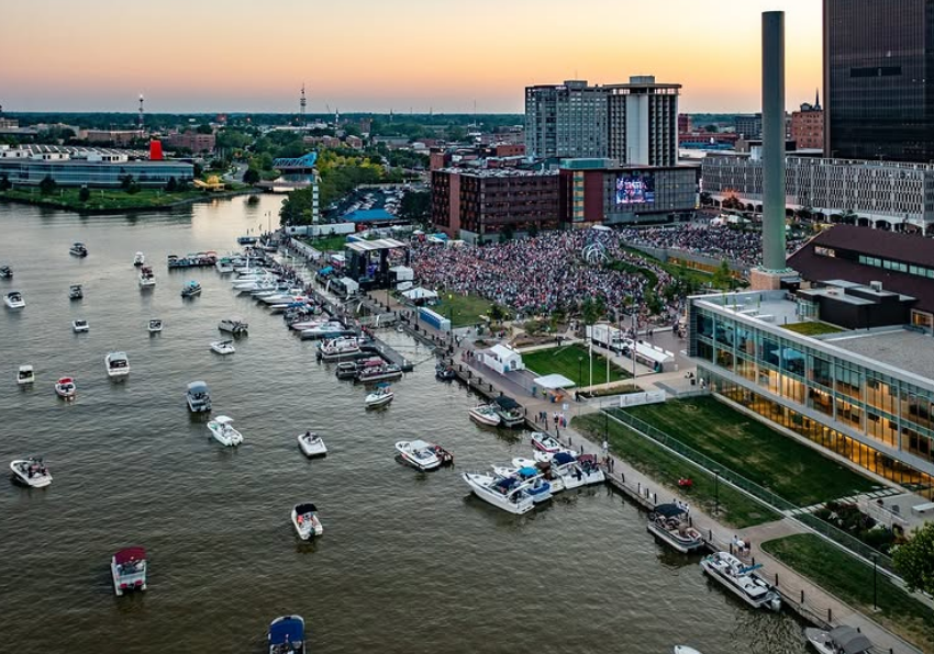 A waterfront cityscape at sunset with boats in the water, a crowd gathered on the shore near a stage, and tall buildings in the background.