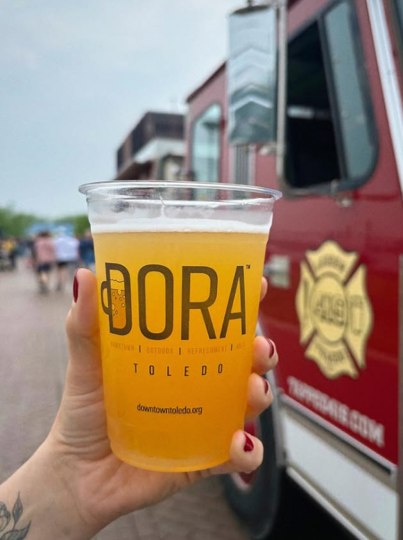 A hand holding a clear plastic cup with a yellow beverage, with the word 'DORA' and 'TOLEDO' printed on it, in front of a red food truck.