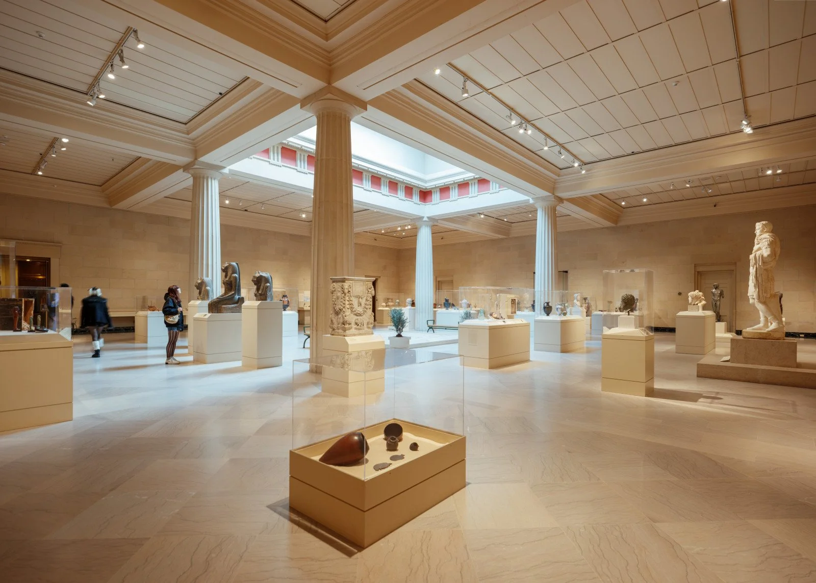 Interior view of a museum gallery with ancient sculptures and artifacts displayed on pedestals and in glass cases, featuring marble columns and a skylight ceiling.